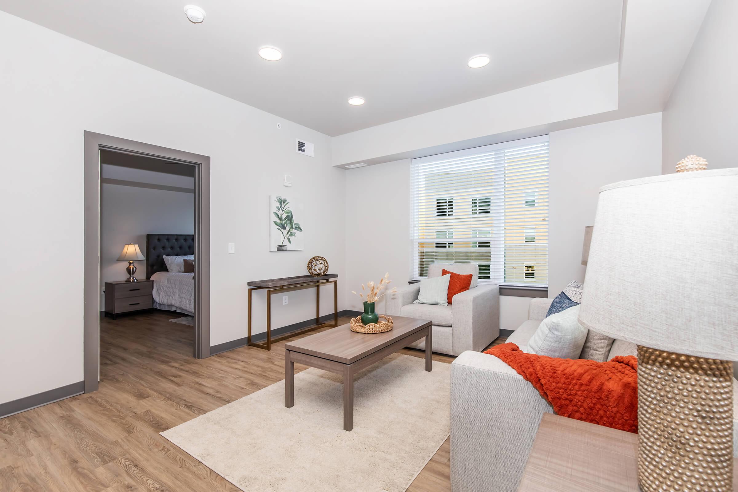 A modern living room with light gray walls and a cozy atmosphere. It features a soft beige sofa with orange cushions, a wooden coffee table, and a floor lamp. A decorative plant and a wall art piece are present. In the background, a doorway leads to a bedroom with light-colored bedding and large windows.