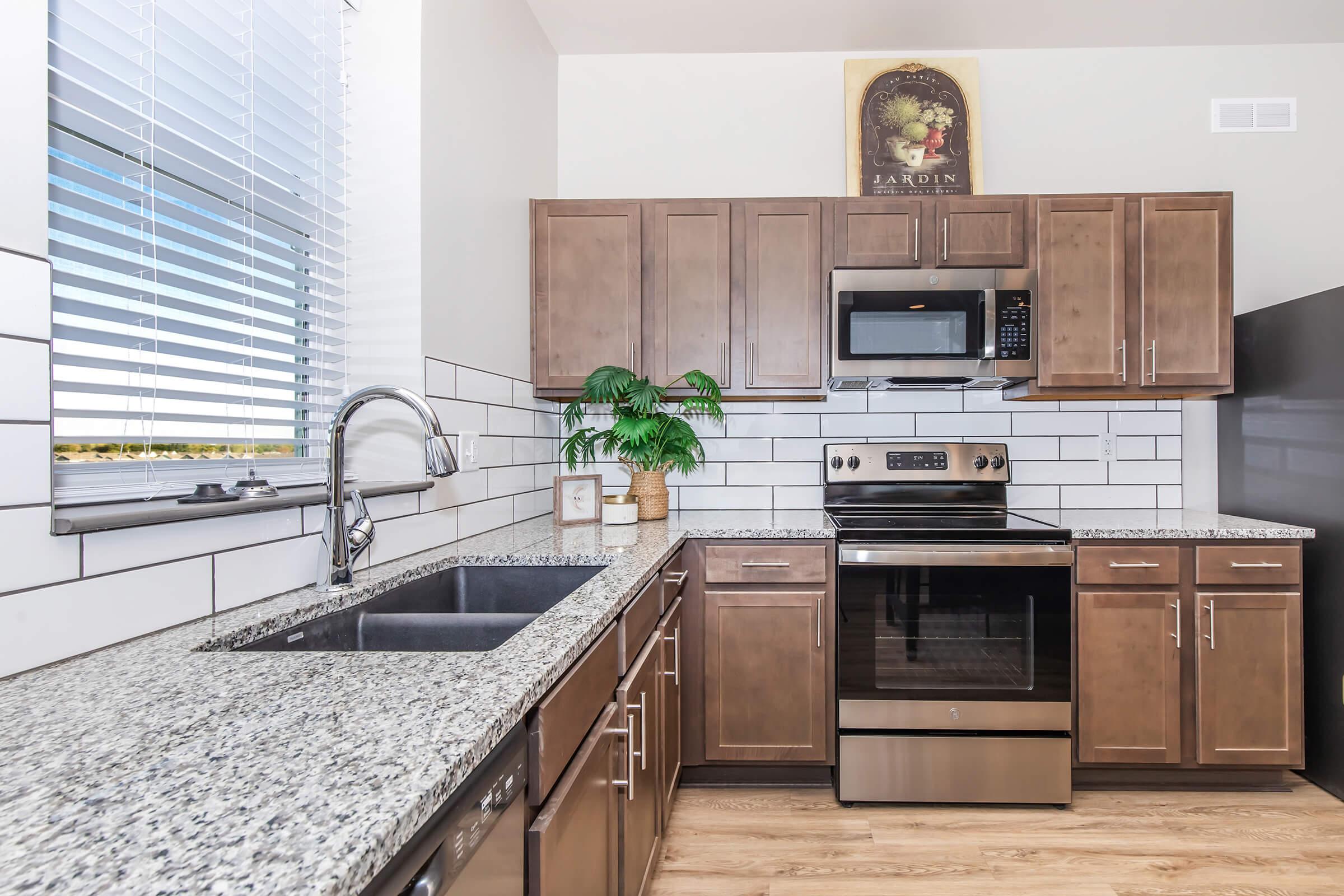 Modern kitchen featuring dark wooden cabinets, a granite countertop, and stainless steel appliances, including a microwave and oven. A potted plant adds a pop of green, while a decorative wall art piece is displayed above the cabinets. Natural light streams in through a window with horizontal blinds.