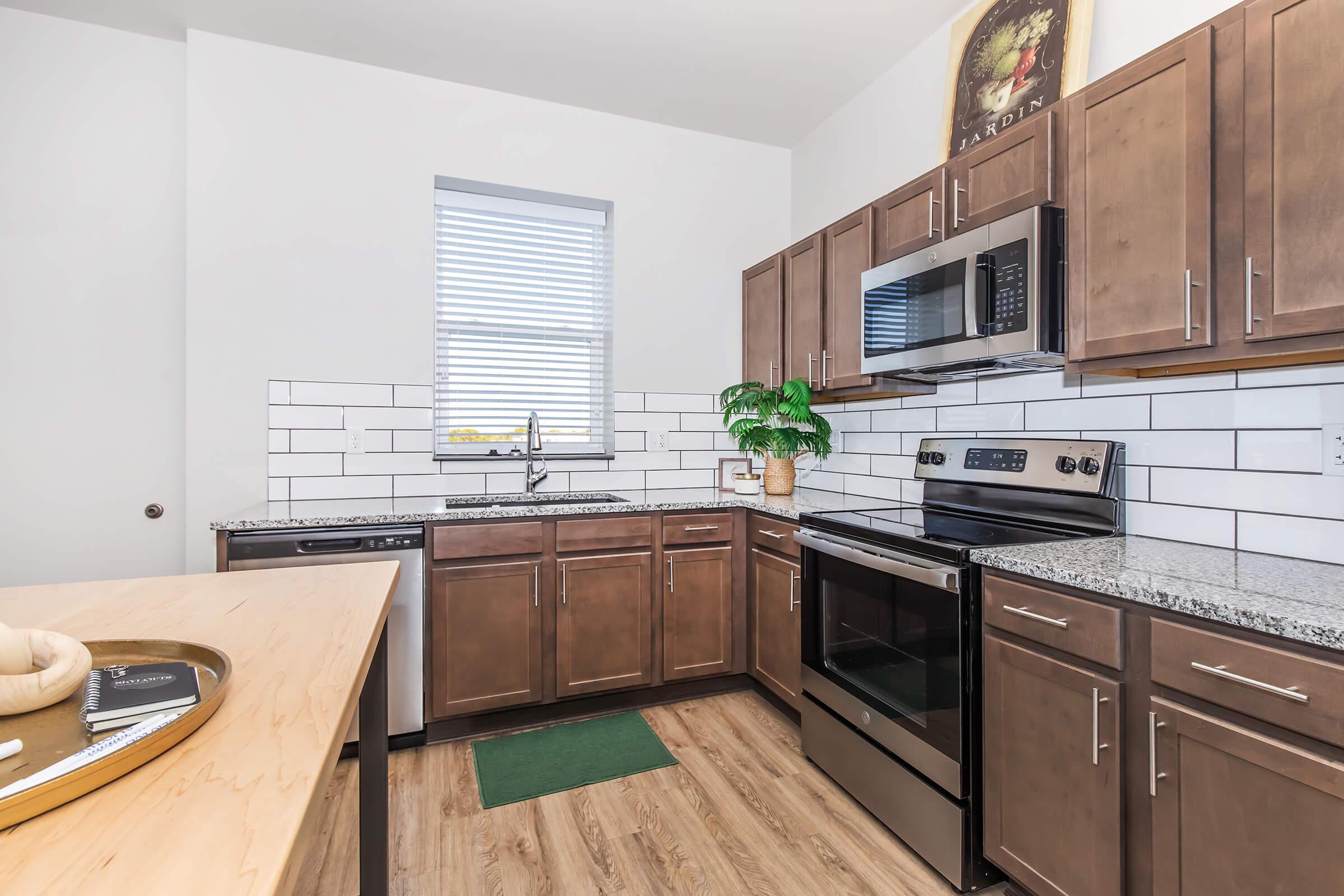 Modern kitchen featuring wooden cabinetry, a stainless steel microwave and oven, and a granite countertop. The space includes a sink with a plant nearby, a window with blinds, and a green mat on the floor. The design is bright and inviting with a contemporary style.