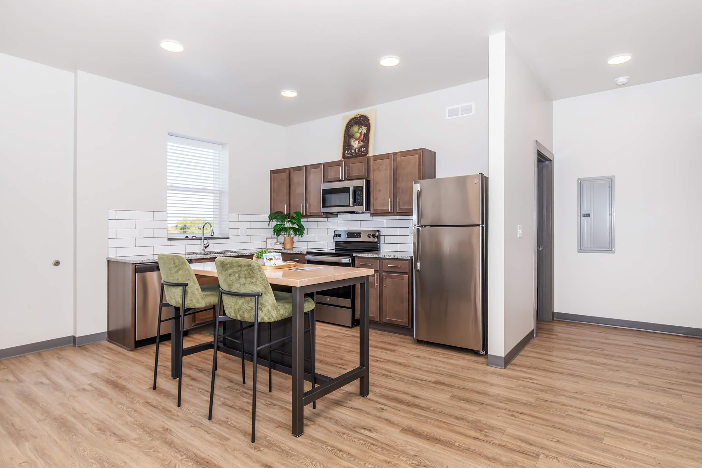 A modern kitchen featuring wooden cabinetry, stainless steel appliances, and a dining table with green upholstered chairs. The flooring is light wood, and the space has ample natural light from a window. A small plant adds a touch of greenery to the decor.