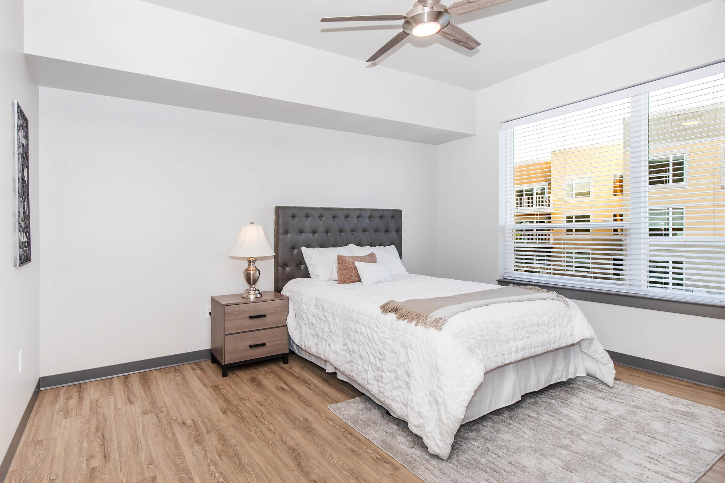 A cozy bedroom with a gray upholstered bed, white bedding, and a lamp on a nightstand. A window with blinds lets in natural light, revealing views of an exterior building. The floor is wooden, and there is a light-colored area rug beneath the bed, creating a warm and inviting atmosphere.