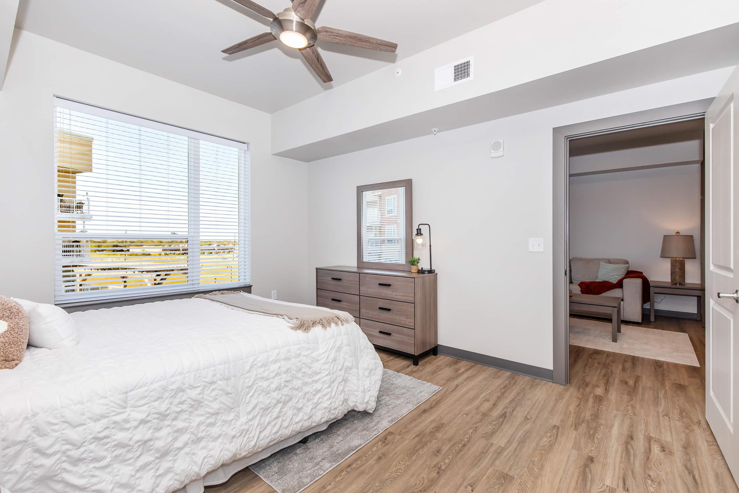 A well-lit bedroom featuring a queen-sized bed with a white quilt, a wooden dresser, and a ceiling fan. There are large windows allowing natural light and an adjoining room visible through an open door, showcasing a cozy sitting area with a light-colored armchair.