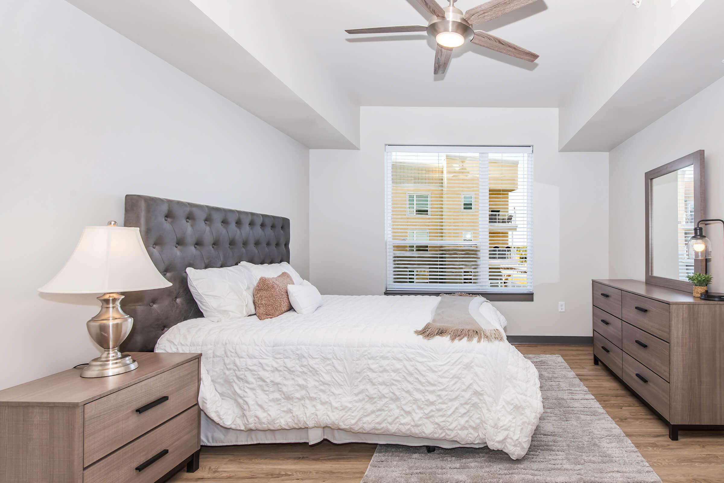 A modern bedroom featuring a tufted gray bed with white bedding, two wooden nightstands with lamps, a dresser, and a large mirror. The room has a window with blinds, allowing natural light to enter, and a light-colored area rug on a wooden floor. The decor is minimal and stylish.