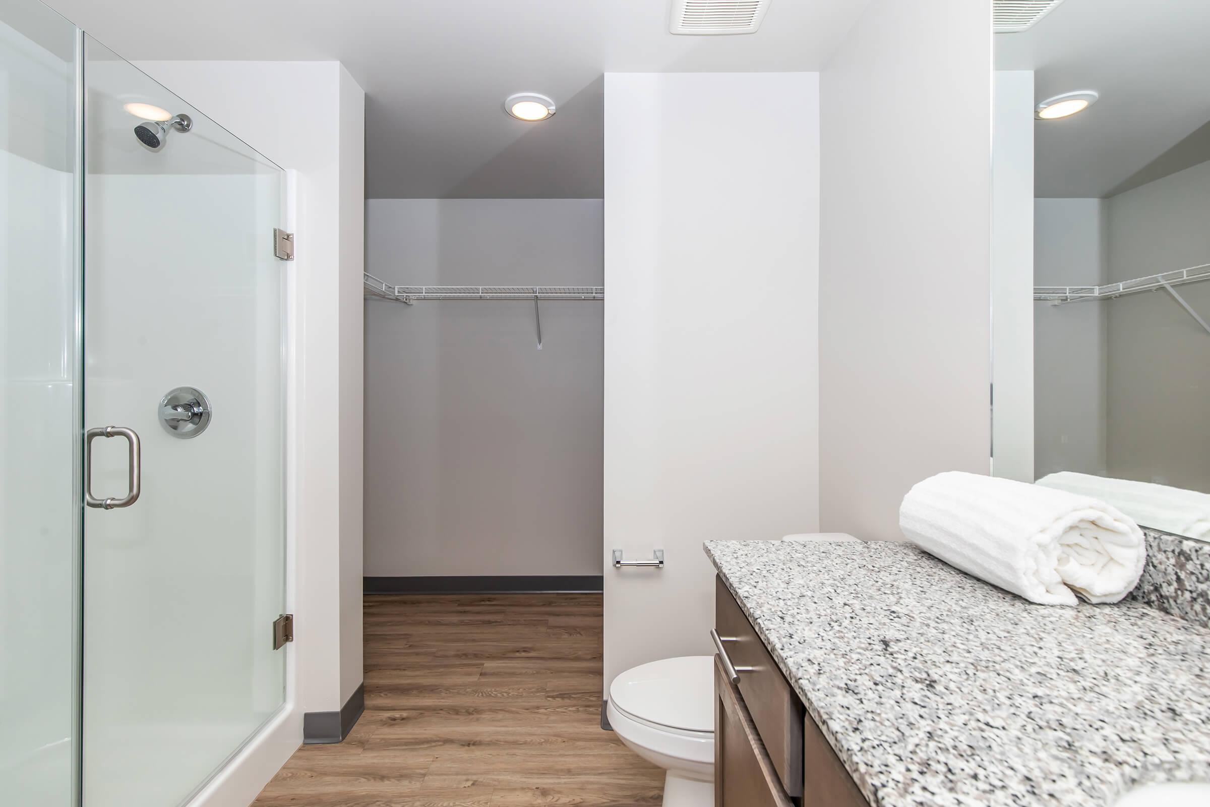 Modern bathroom featuring a glass shower, a clean toilet, and a granite countertop. A neatly rolled white towel is placed on the counter, and the space includes sleek wooden flooring and a towel rack in the background. The design emphasizes simplicity and functionality with neutral colors.