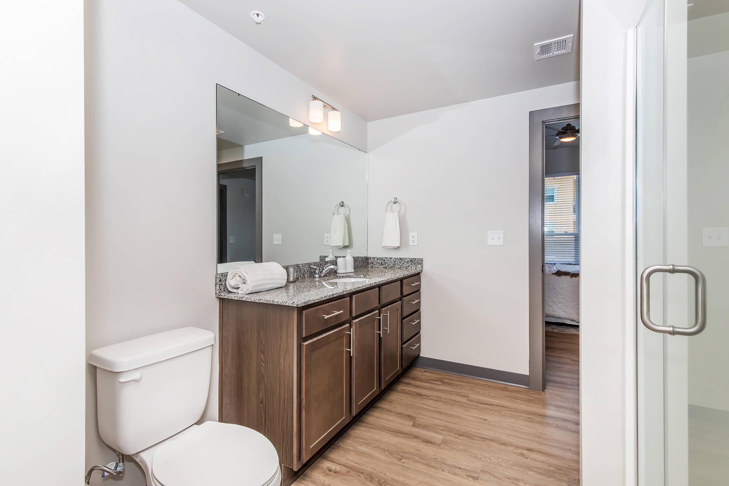 A modern bathroom featuring a granite countertop with dark wood cabinets, a large mirror above the sink, and white towels hanging nearby. The floor is hardwood, and a shower with a glass door is visible. The bathroom design is sleek and contemporary, with neutral wall colors and good lighting.