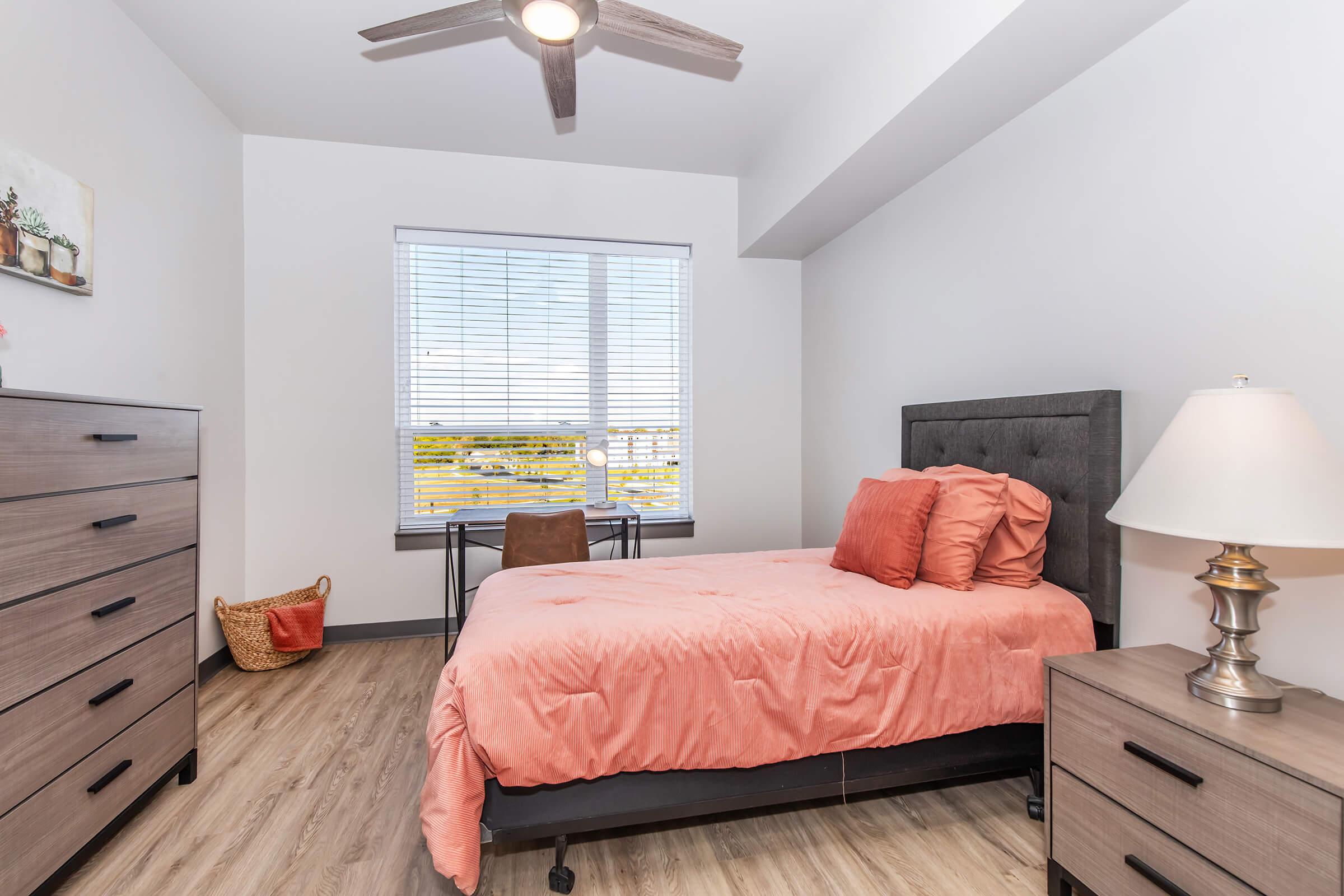 A bright, modern bedroom featuring a gray upholstered bed with coral bedding, a nightstand with a lamp, a wooden dresser, and a small workspace with a chair. Natural light floods the room through a large window with white blinds, showcasing a view of the outdoors.