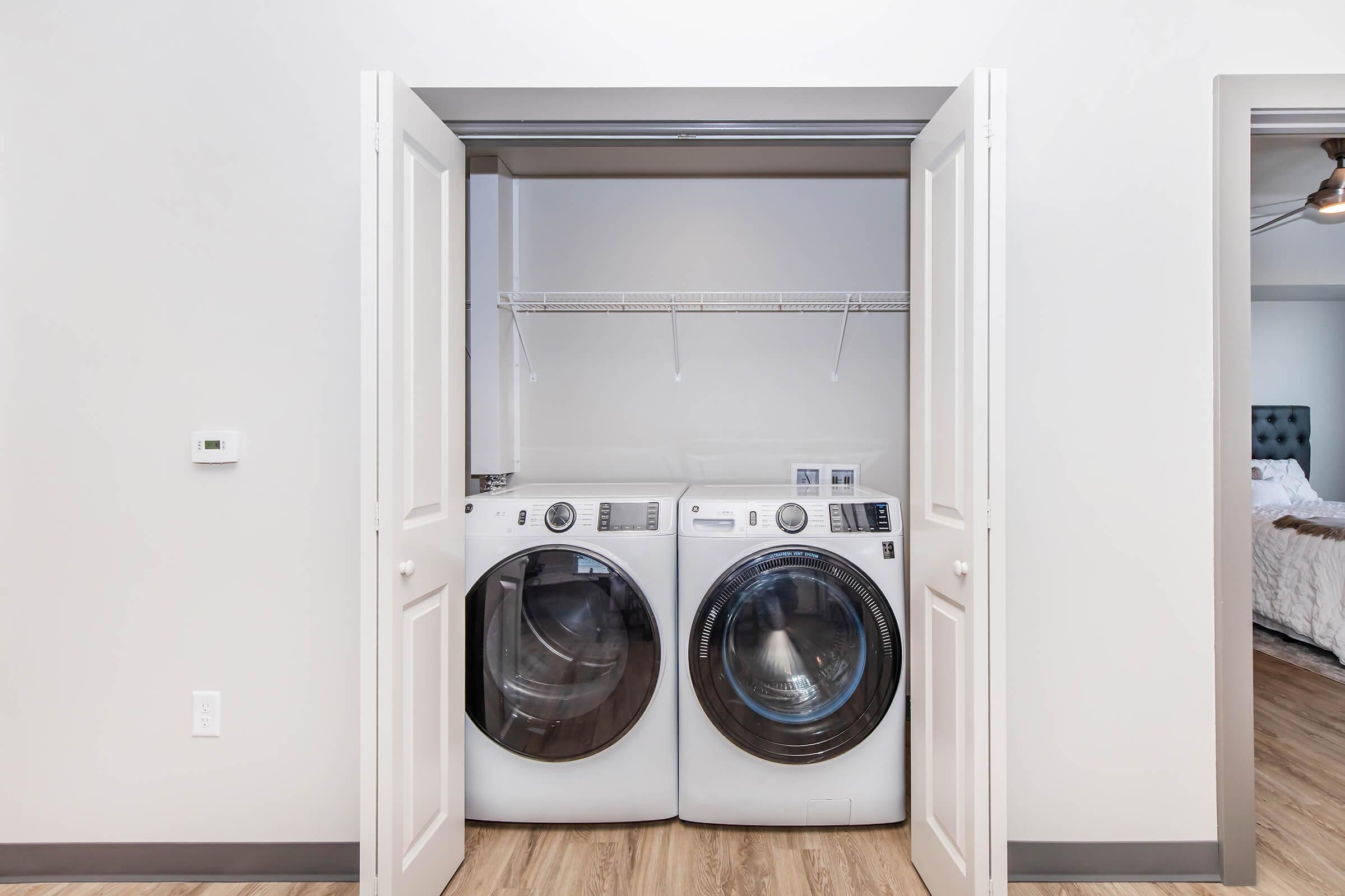 A laundry closet featuring a stacked washer and dryer set in white. The doors are open, revealing the appliances on a shelf. The space has light-colored walls and a wooden floor, creating a clean and organized appearance.
