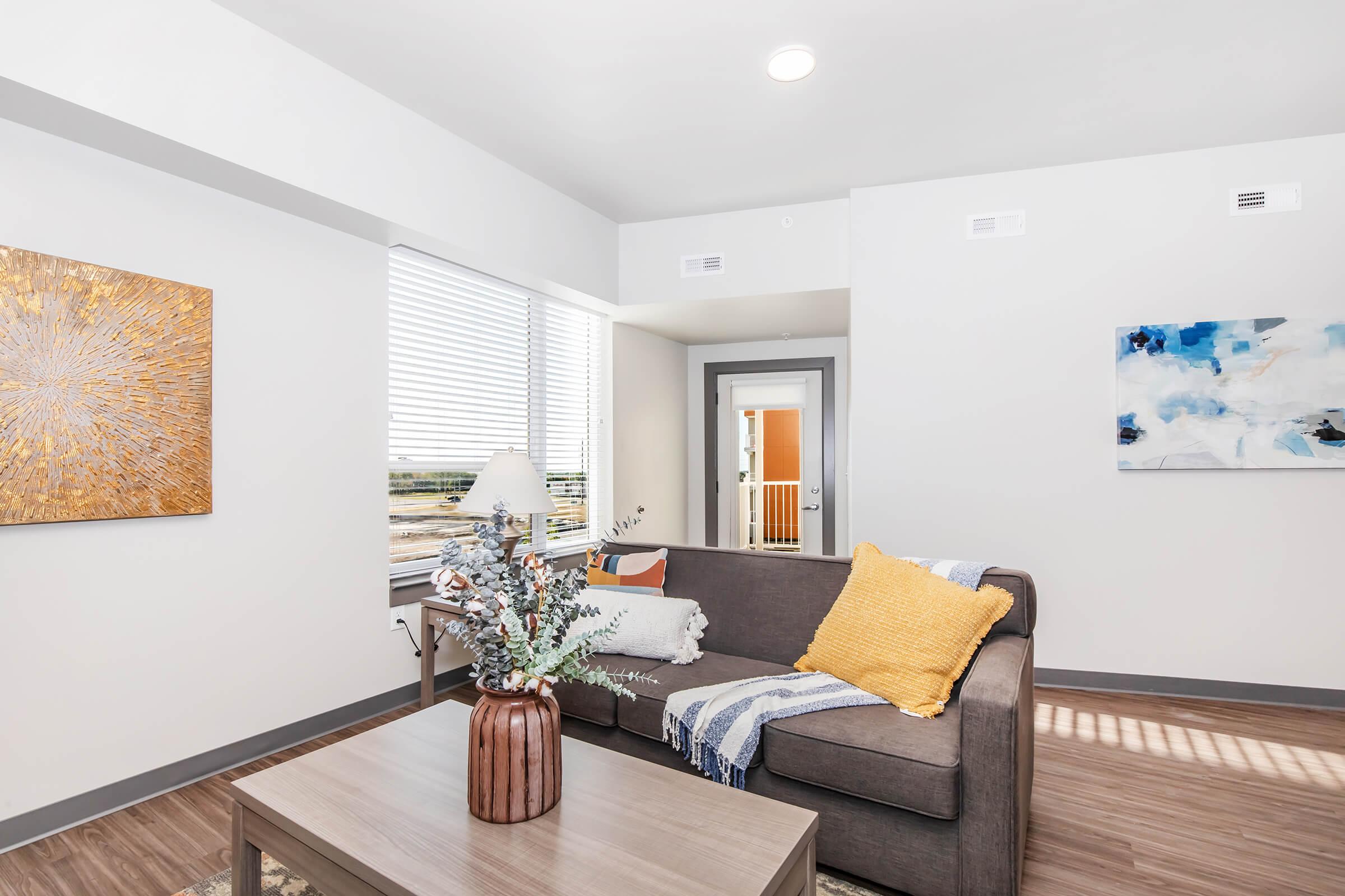 A modern living room featuring a brown couch with a yellow throw pillow, a coffee table with decorative flowers, and two pieces of abstract wall art. Large windows let in natural light, and a door leads outside. The flooring is laminate, and the decor includes a blue and white striped blanket.