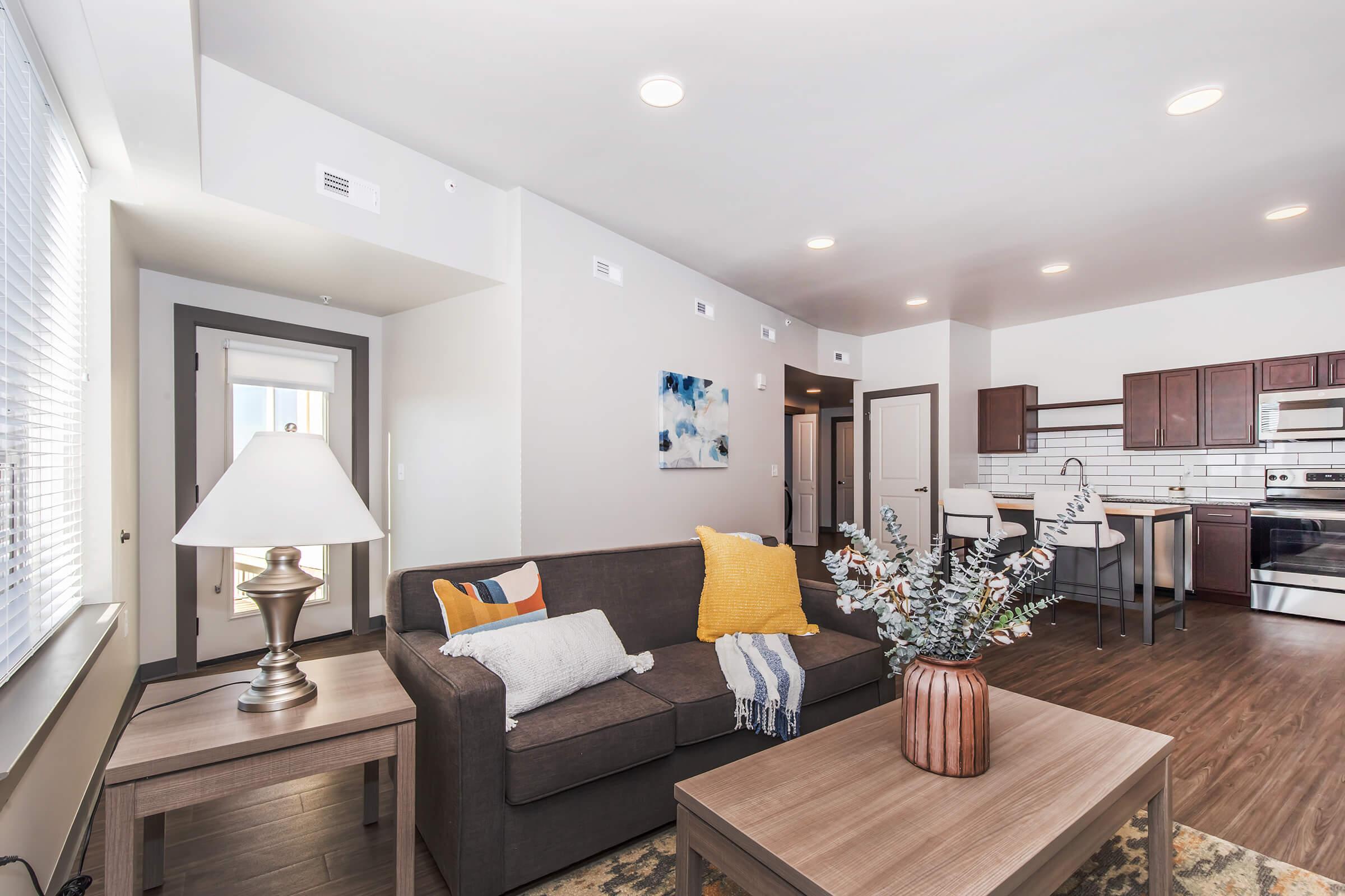 A modern living room featuring a beige sofa with colorful cushions, a wooden coffee table, and a lamp. Natural light fills the space from large windows. The background includes a dining area with a small table and chairs, along with a contemporary kitchen visible, equipped with stainless steel appliances.