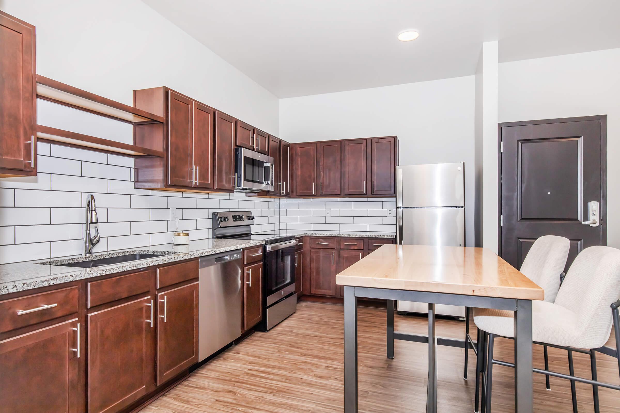 Modern kitchen featuring dark wood cabinetry, stainless steel appliances, and a spacious countertop. There is a breakfast bar with high chairs, and the walls are adorned with white subway tiles. The flooring is a light wood finish, creating a warm and inviting atmosphere.