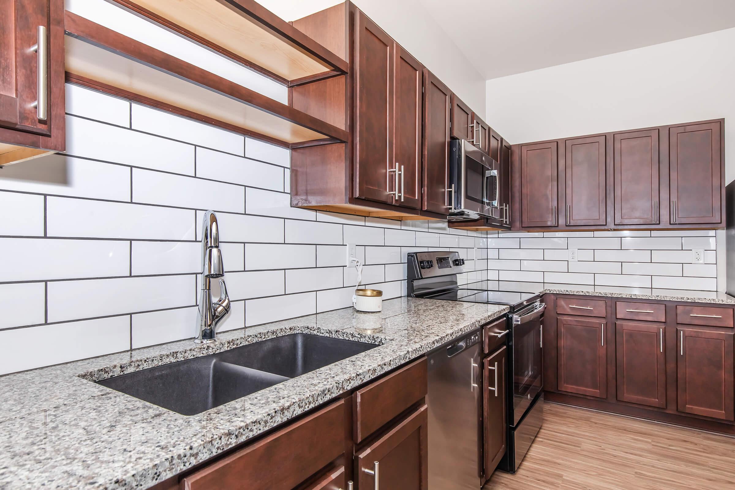 Modern kitchen featuring dark wood cabinets, a stainless steel sink, and a granite countertop. The backsplash consists of white subway tiles, and there is a stainless steel range and microwave. Open shelving above the countertop provides storage space. The flooring is a light wood laminate.