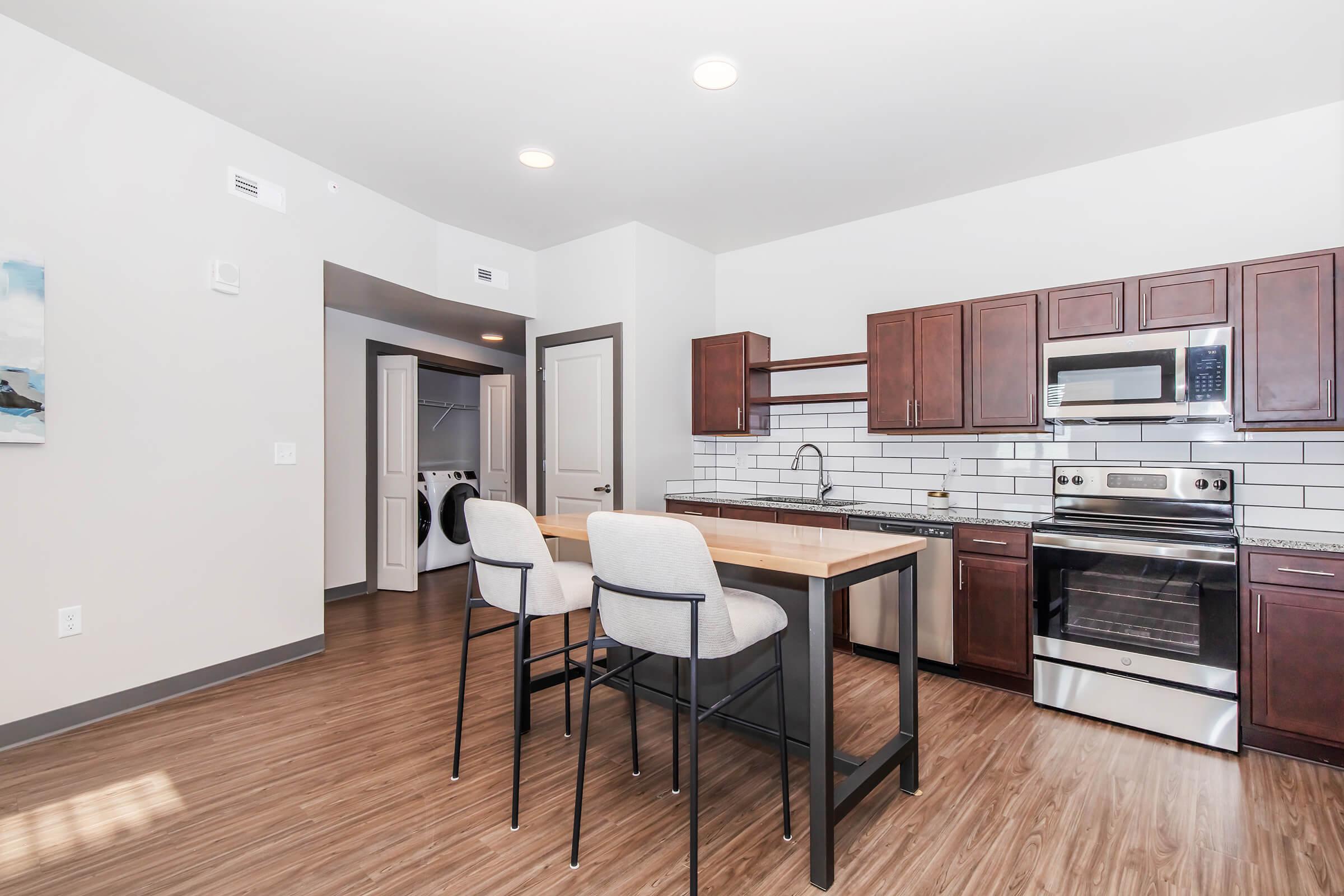 Modern kitchen with dark wooden cabinetry, stainless steel appliances, and a large island with two barstools. Features a tiled backsplash and an open layout towards a laundry area with a door. The flooring is light wood, and the room is bright with overhead lighting.
