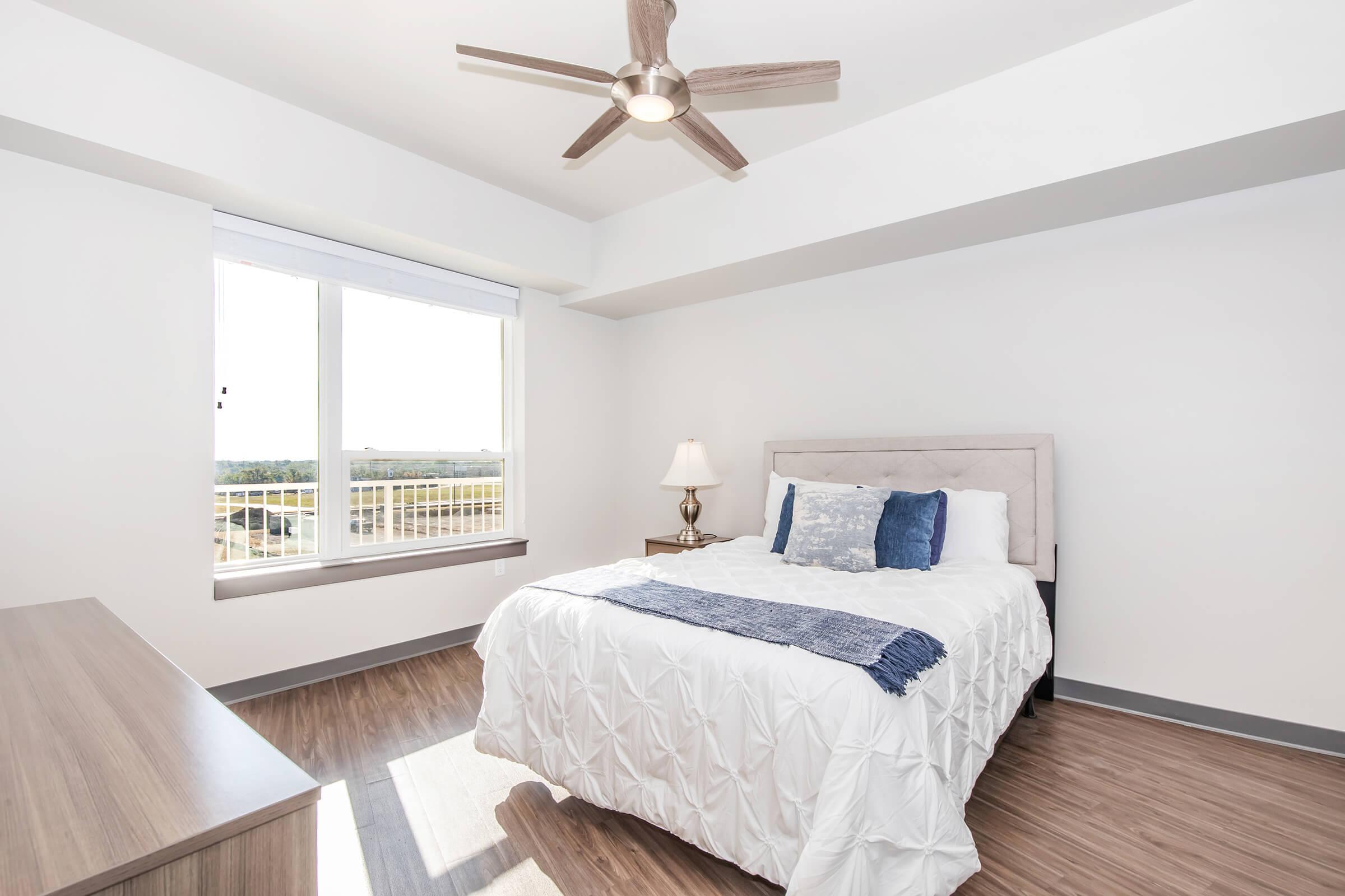 A bright and modern bedroom featuring a queen-sized bed with white linens and blue accent pillows. There’s a nightstand with a lamp beside the bed, a dresser against the wall, and large windows providing natural light and a view of the outdoors. The flooring is wood-like, adding warmth to the space.