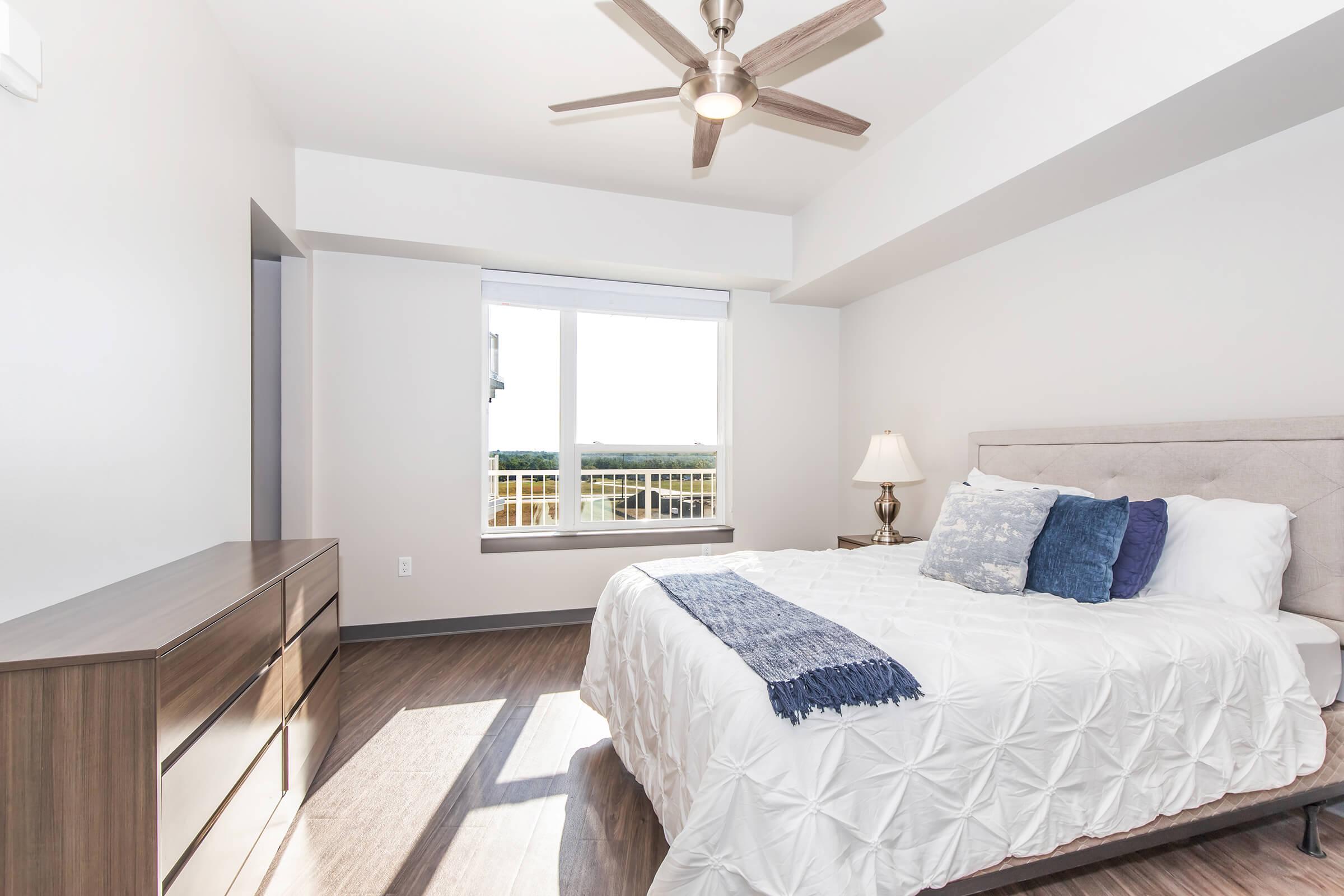 A modern bedroom featuring a queen-sized bed with a light grey upholstered headboard and decorative pillows. There is a nightstand with a lamp beside the bed, a brown dresser against the wall, and a large window providing natural light and a view outside. The room has light-colored walls and wooden flooring.