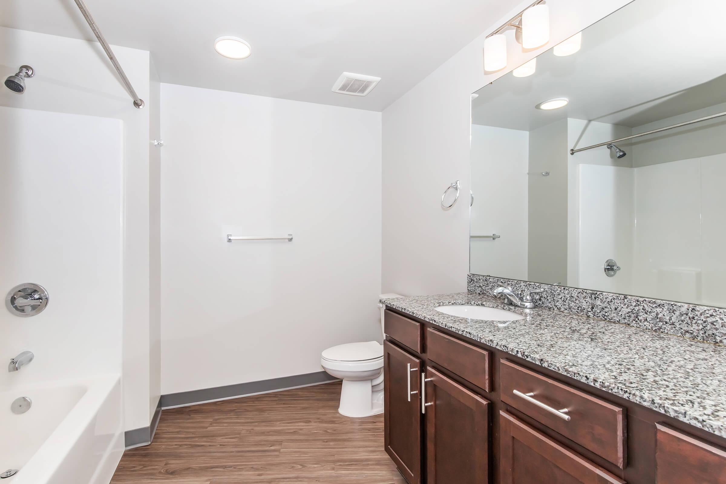 A modern bathroom featuring a white bathtub with a shower, a glass shower wall, a large mirror above a dark wood vanity with a granite countertop, and a white toilet. The floor is a wood-like laminate, and the walls are painted white, creating a clean and bright atmosphere.