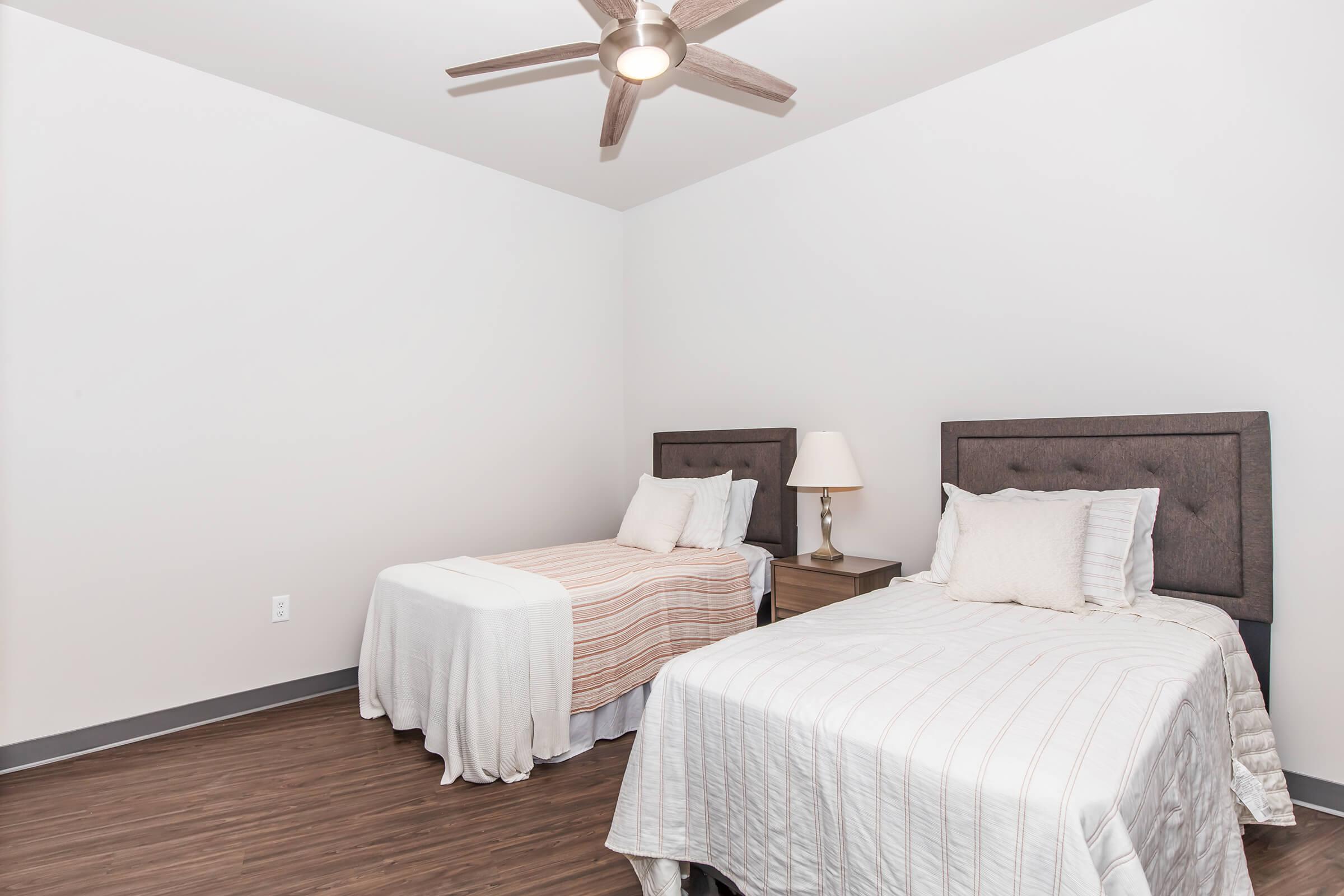 A neat and modern bedroom featuring two twin beds with light-colored bedspreads. Each bed is paired with a dark wood nightstand, one lamp, and there is a ceiling fan above. The walls are painted white, and the floor has a wooden appearance, creating a clean and simple aesthetic.