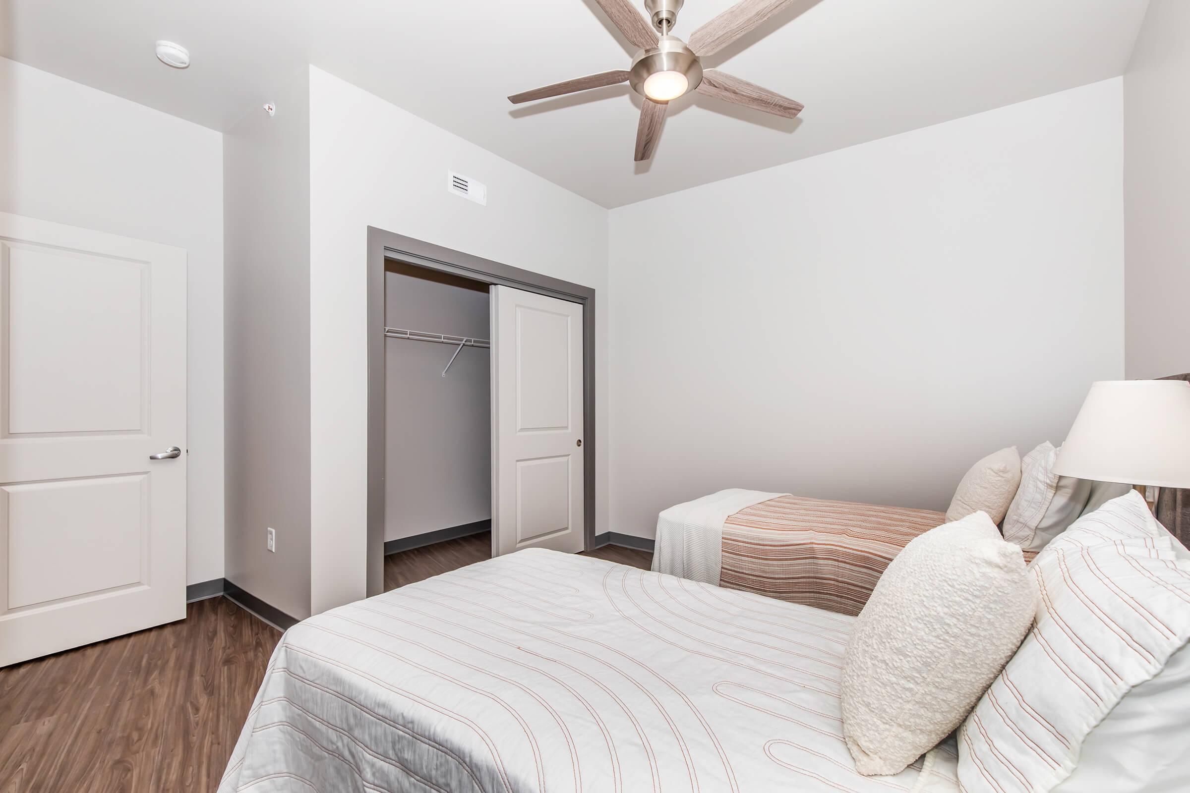 A bright bedroom featuring two twin beds with light-colored bedding. A ceiling fan is visible above. There's a closet area with an open door, and the walls are painted in a soft neutral color, complementing the contemporary design of the space. The floor is a modern wood-like finish.