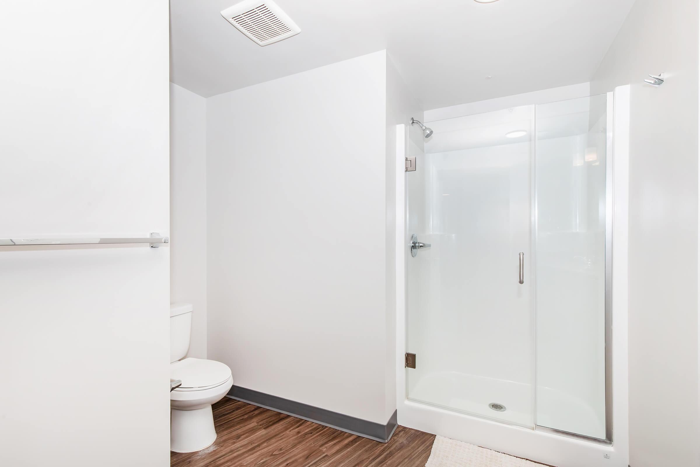 A modern bathroom featuring a glass shower enclosure, a toilet, and light-colored walls. The floor is made of wood-like material, and a small rug is placed near the shower. The space is well-lit, creating a clean and spacious appearance.