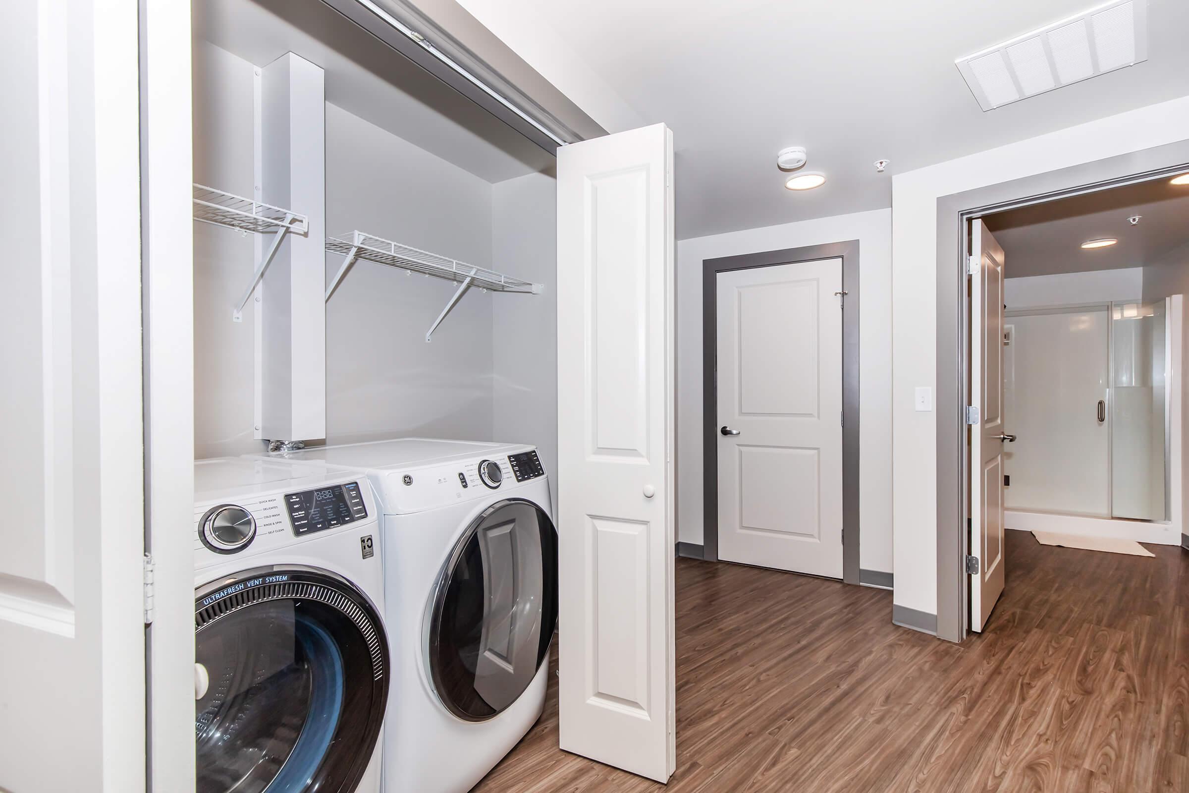 A clean and modern laundry room featuring a stacked washer and dryer, with a white shelving unit above. The room has neutral walls and a wood-like floor, along with a door leading to another space, indicating an organized and efficient laundry area in a residential setting.