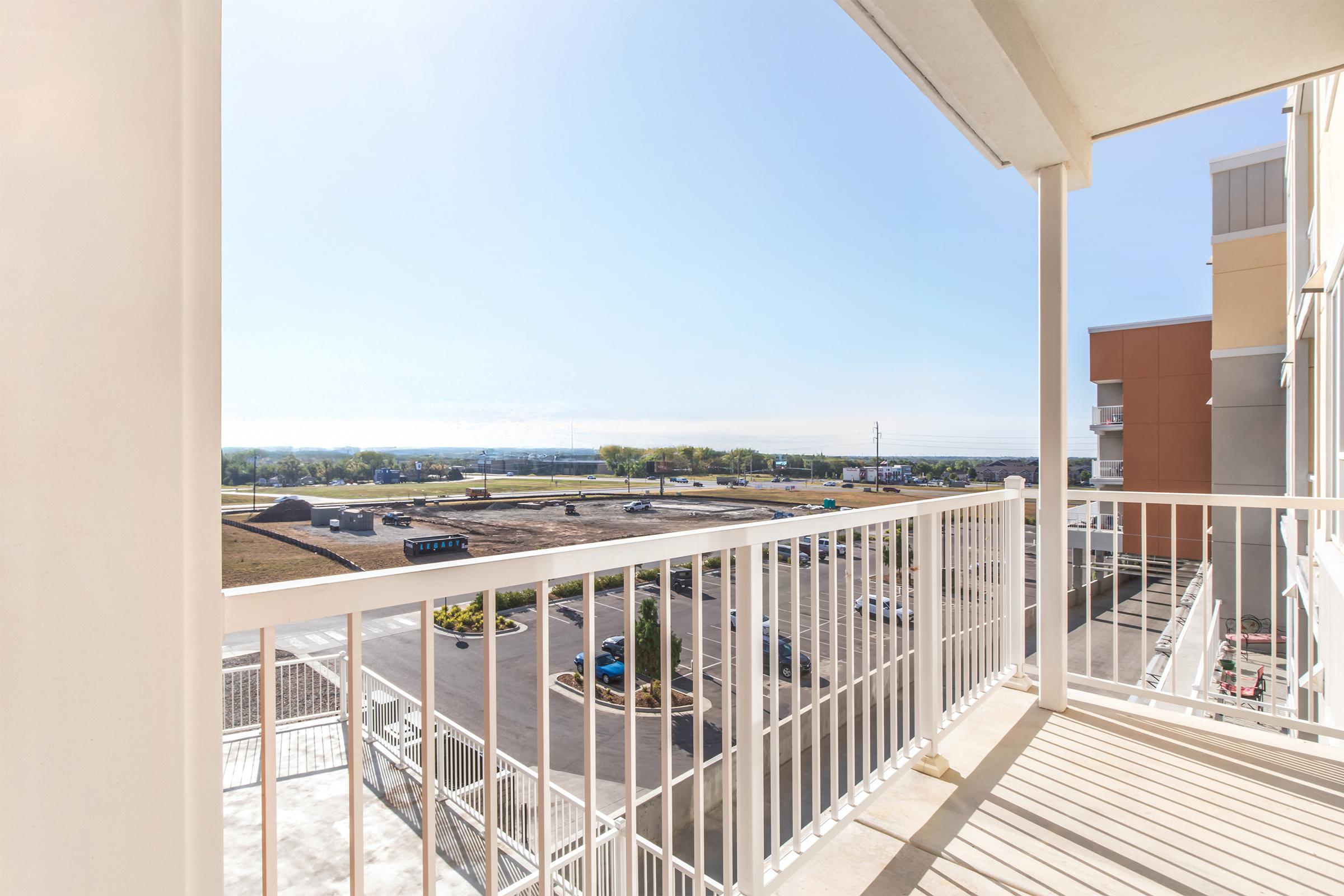 A view from a balcony overlooking a parking area and an expansive landscape with greenery, under a clear blue sky. The sun is shining brightly, creating a warm, inviting atmosphere. The building's exterior features light-colored walls and a white railing.