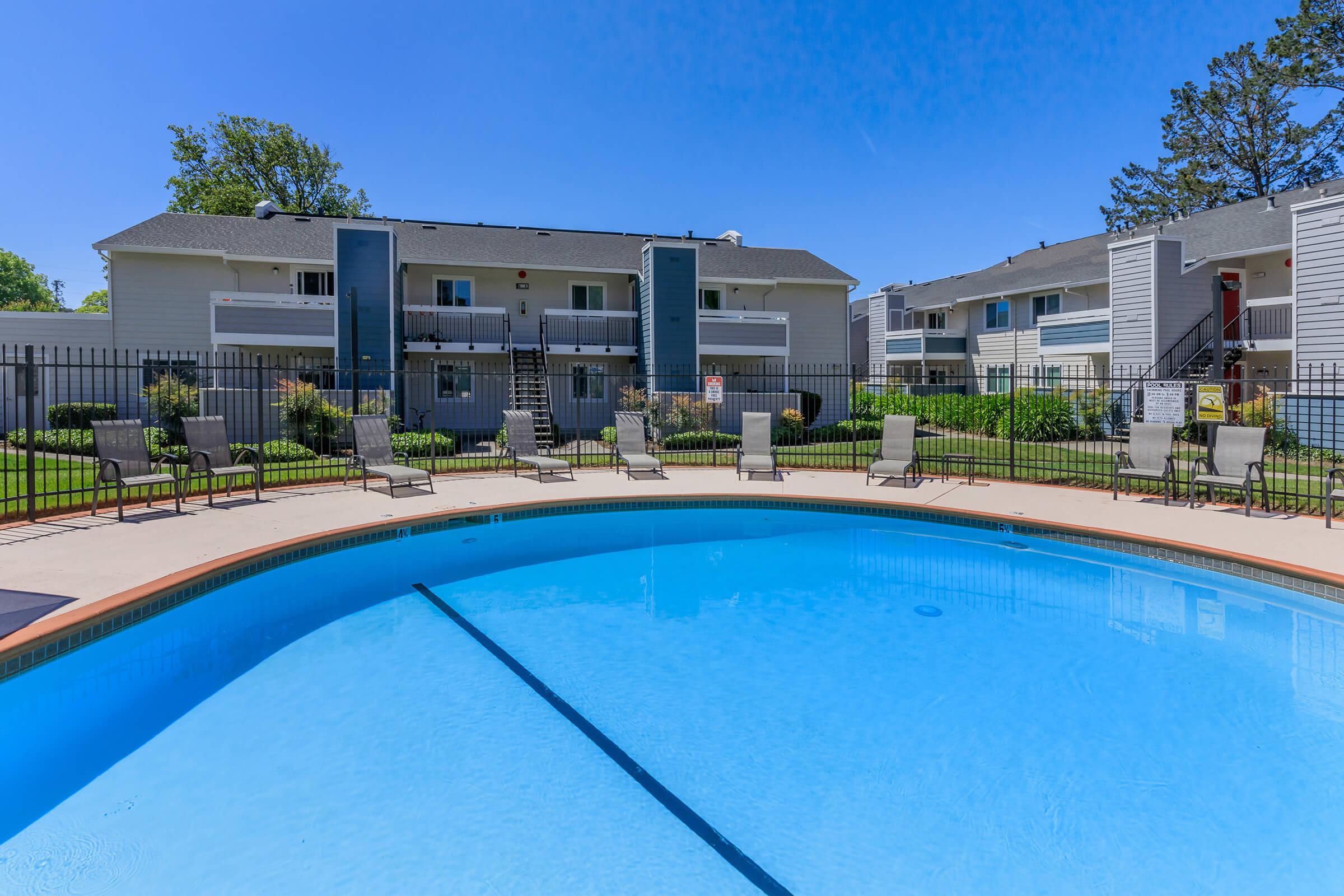 A clear blue swimming pool surrounded by lounge chairs, with a fenced lawn area and two-story apartment buildings in the background. The sky is clear and blue, creating a bright and inviting atmosphere.