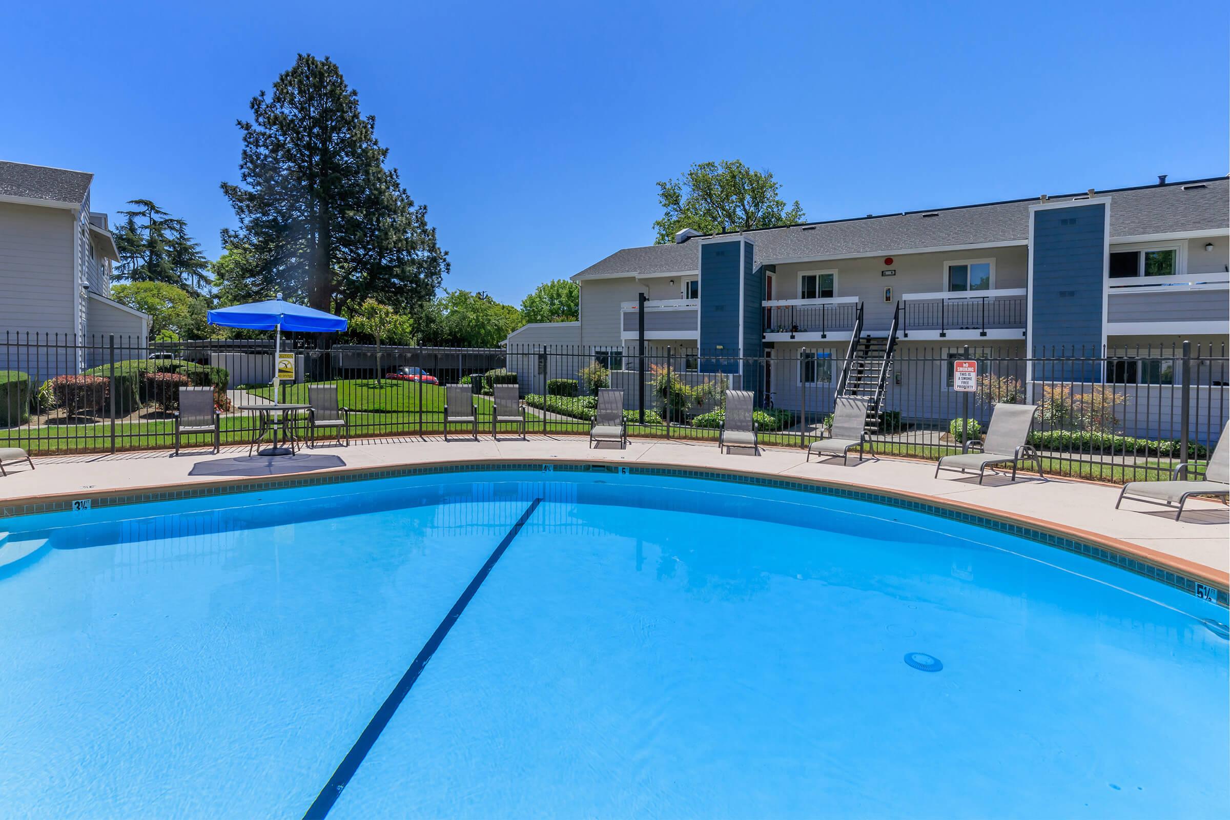 A swimming pool with clear blue water, surrounded by lounge chairs and a fence. In the background, there are two-story buildings and green landscaping under a bright blue sky. A large umbrella is set up near the pool, providing shade.
