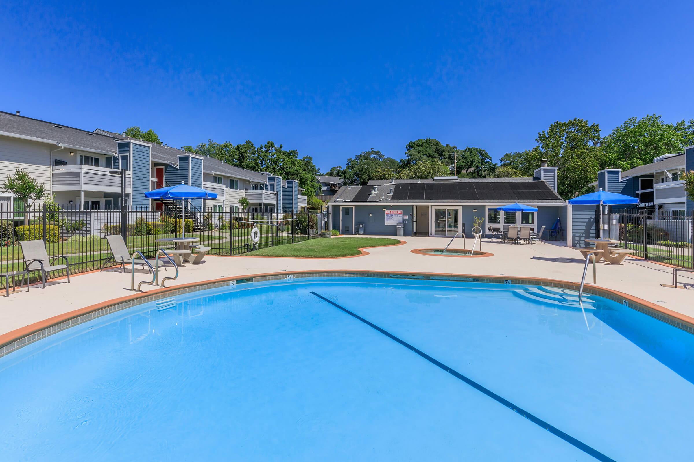 A clear blue swimming pool surrounded by lounge chairs and shaded umbrellas. In the background, there are modern apartment buildings and a well-maintained lawn, with a bright blue sky overhead. The scene conveys a relaxing outdoor space ideal for leisure and recreation.