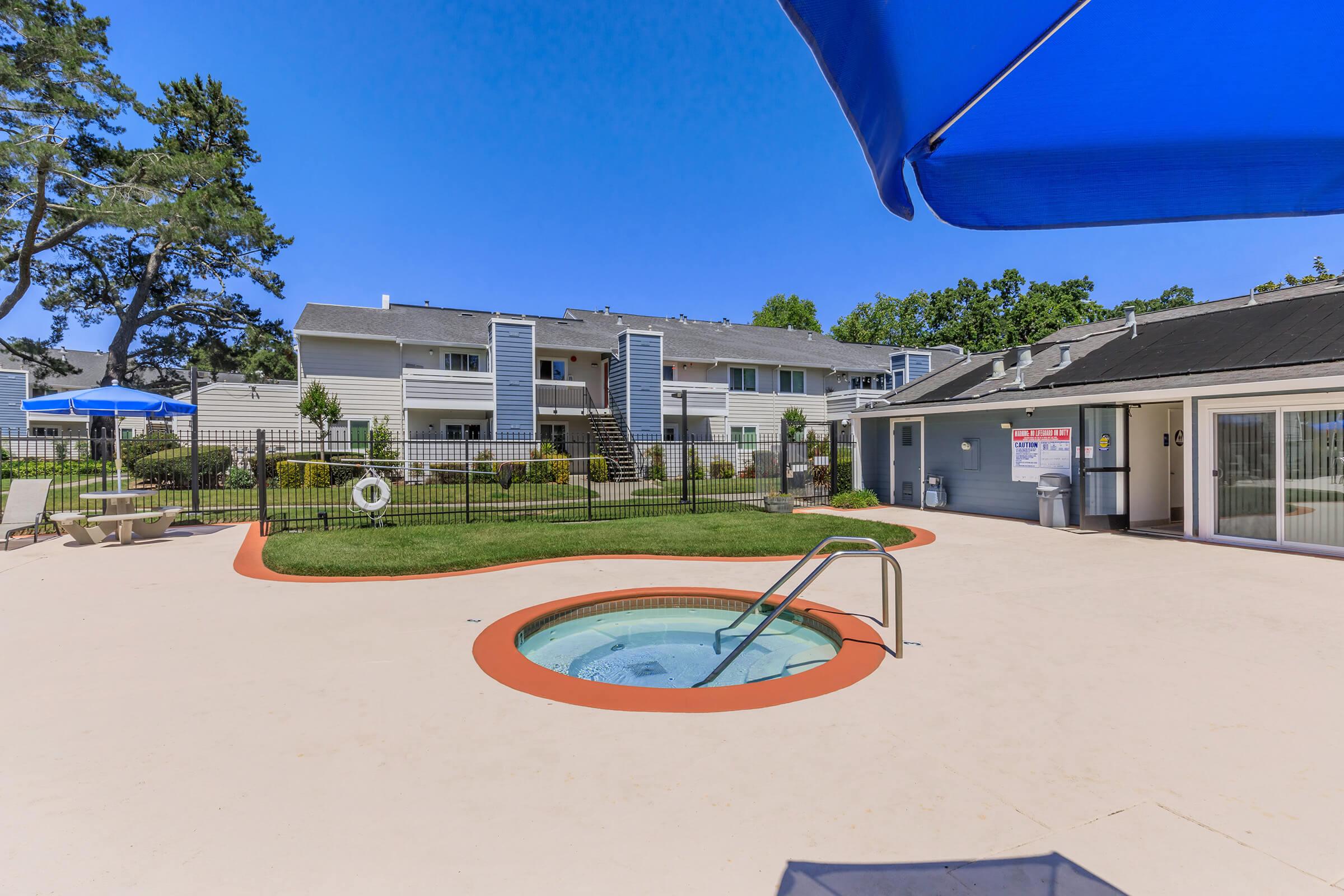 A sunny outdoor area featuring a small hot tub surrounded by a paved surface. In the background are multi-story residential buildings with green landscaping, umbrellas, and a swimming pool area, all under a clear blue sky. The scene conveys a relaxing atmosphere.