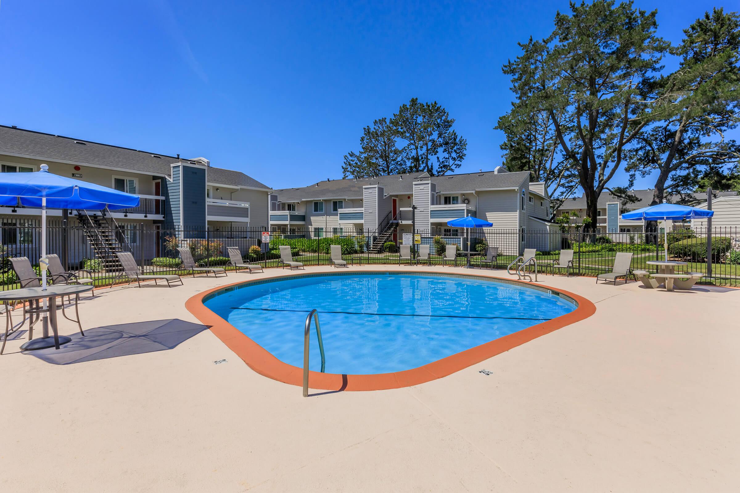 Swimming pool surrounded by lounge chairs and umbrellas, situated in a well-maintained courtyard. In the background, there are two-story apartment buildings with grassy areas and trees under a clear blue sky. The scene conveys a relaxing and inviting outdoor space.