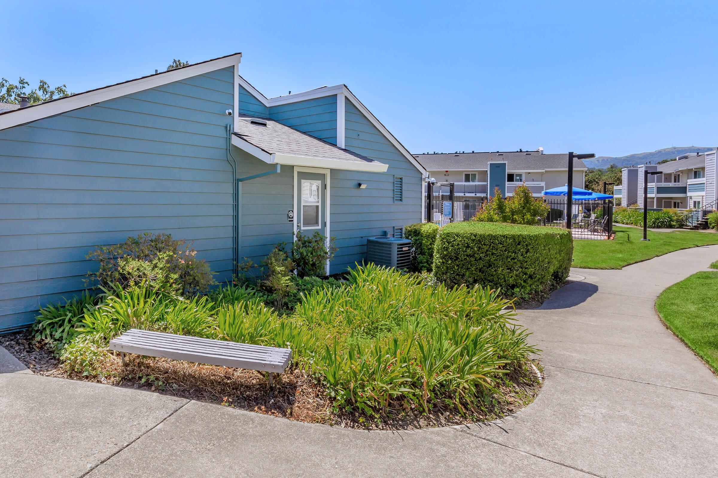 A well-maintained residential area featuring a blue house with a sloped roof, surrounded by lush greenery and ornamental plants. A concrete walkway leads past a bench, with a view of a swimming pool and additional buildings in the background under a clear blue sky.