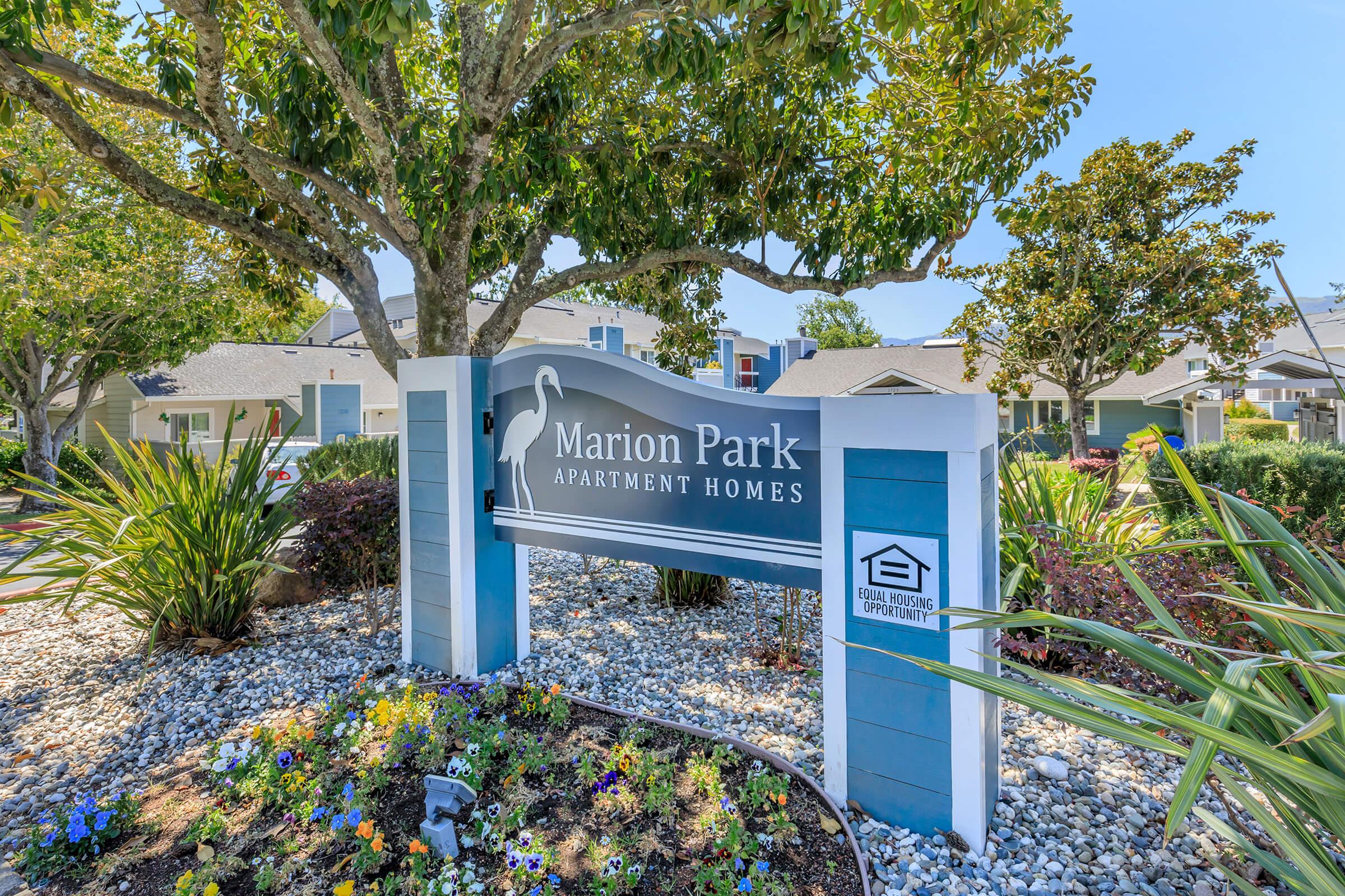 A well-maintained sign for Marion Park Apartment Homes, surrounded by landscaped greenery and colorful flowers. The sign is prominently featured near trees, indicating a welcoming entrance to the apartment complex. Bright blue skies and residential buildings are visible in the background.