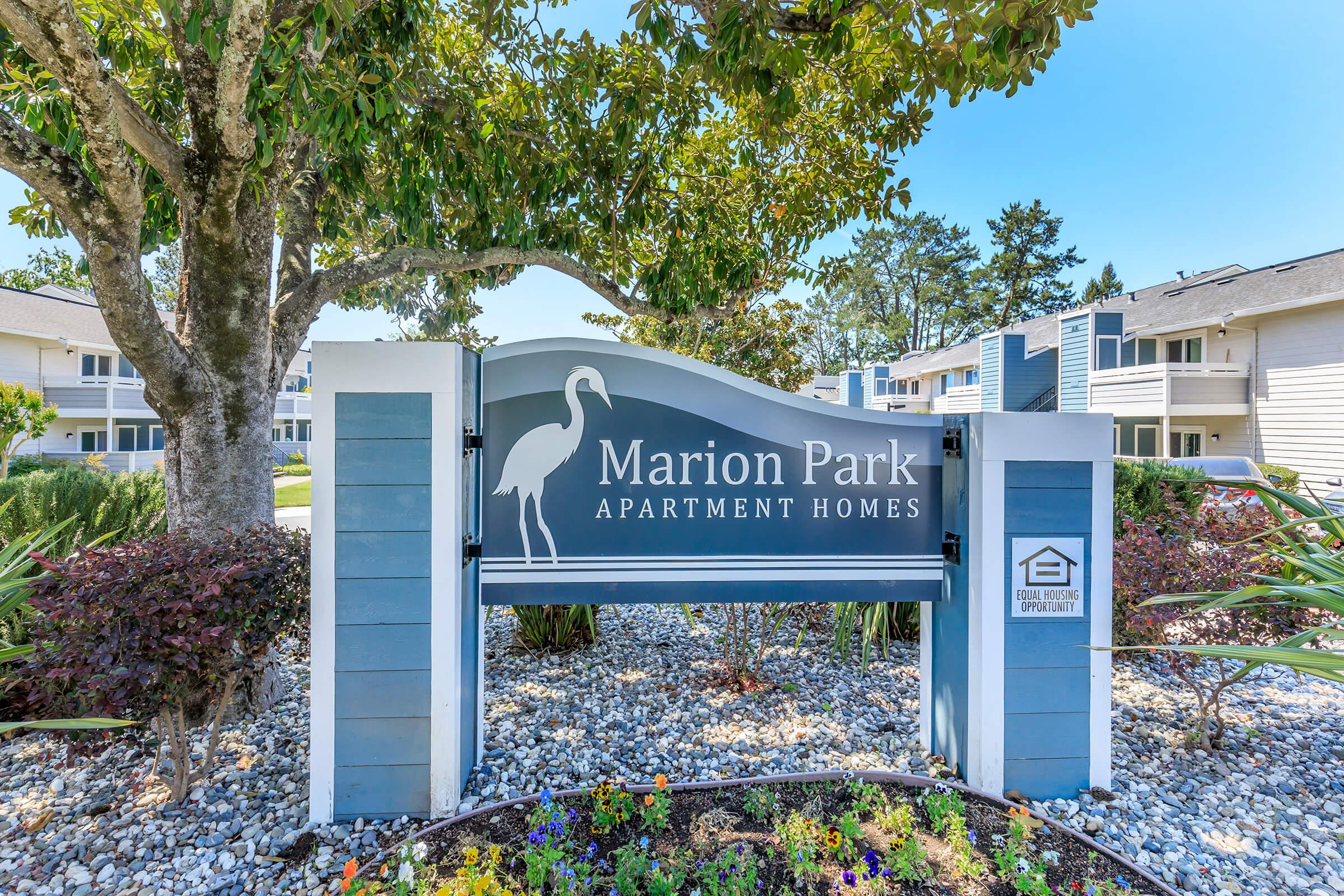 Sign for Marion Park Apartment Homes featuring a silhouette of a heron. The sign is surrounded by well-maintained landscaping with various plants and flowers in bloom. In the background, there are residential buildings and trees under a clear blue sky.