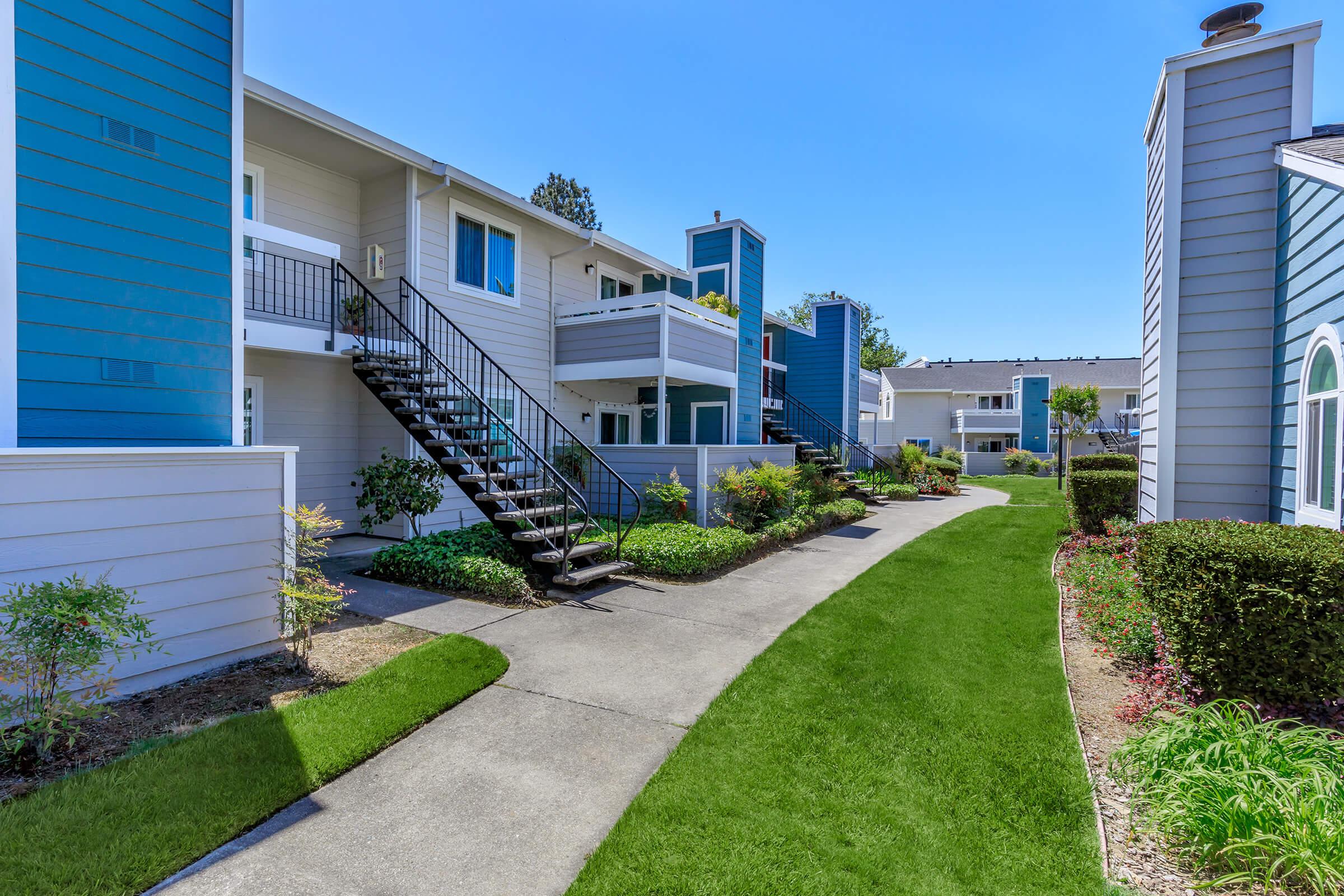 A well-maintained residential complex featuring two-story buildings with blue and gray siding. The pathway is lined with green grass and small shrubs, leading past staircases to the entryways of the apartments. Bright blue skies and a well-kept environment create a welcoming atmosphere.