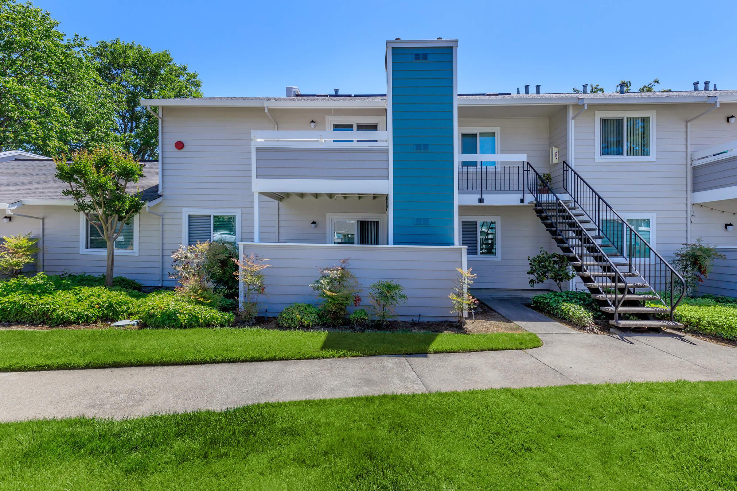 A modern two-story apartment building with light gray siding and a blue accent on the second floor. There are staircases leading to upper units, surrounded by well-maintained green lawns and small shrubs. The sky is clear and blue, adding to the vibrant look of the residence.