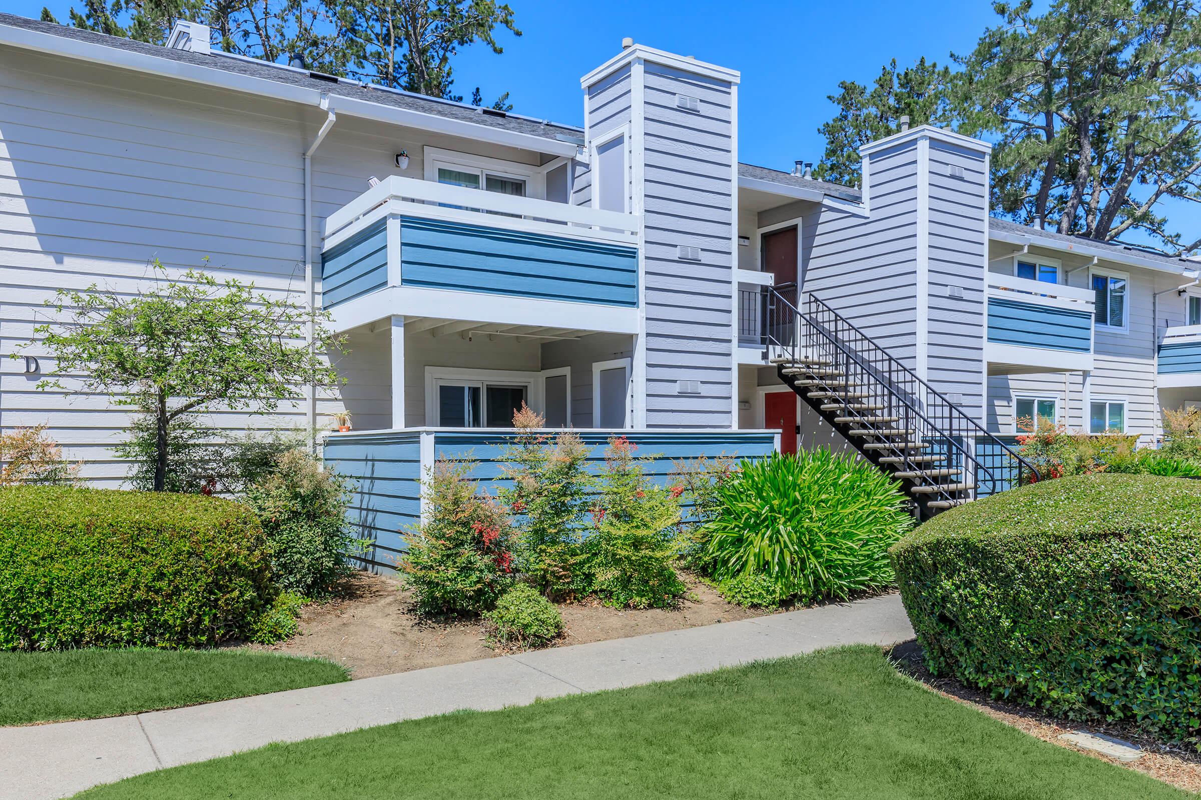 A modern multi-unit residential building with blue and white siding. It features multiple balconies, a staircase, and landscaped greenery, including bushes and plants. The setting is bright and sunny, with clear blue skies and well-maintained surroundings.