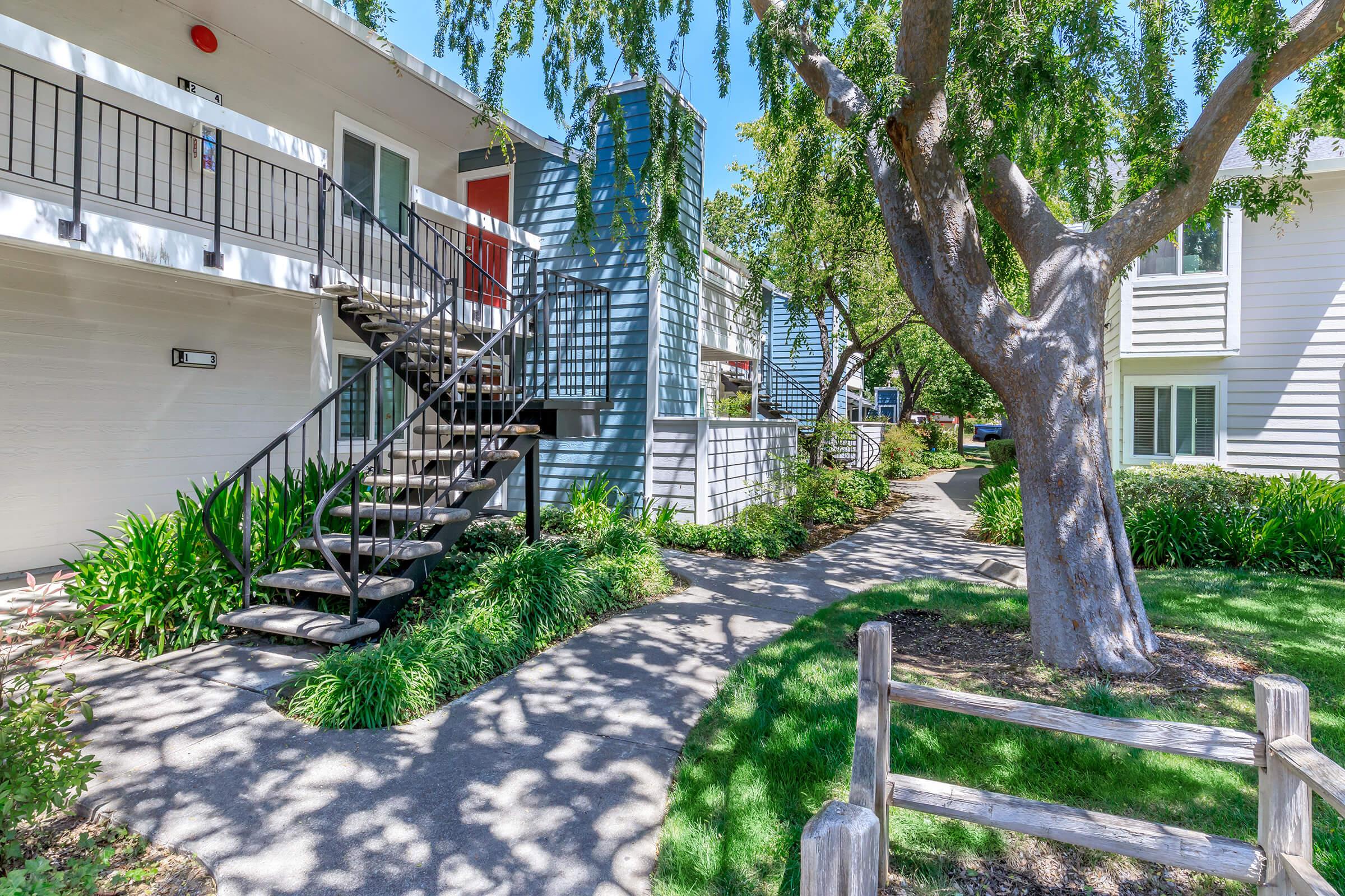 A well-maintained residential complex featuring a winding pathway surrounded by greenery. The path leads past two buildings with balconies and a distinctive blue structure. A large tree provides shade, enhancing the inviting atmosphere of the community.