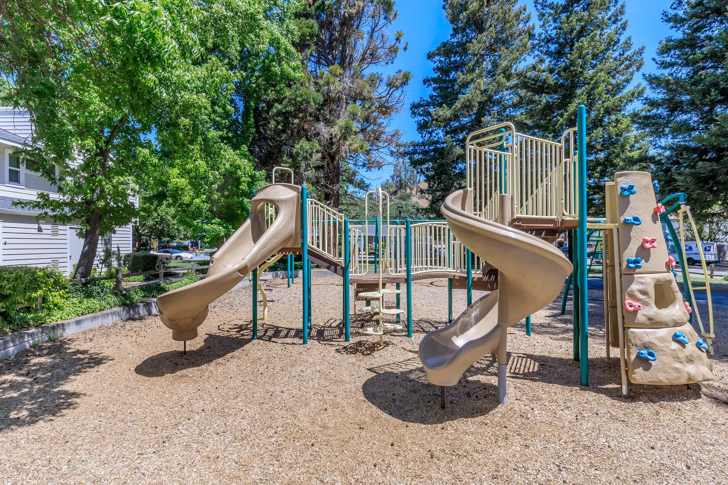 Playground scene with two tan slides, a climbing wall, and various play equipment surrounded by greenery. The ground is covered in wood chips, and tall trees provide shade, creating a sunny outdoor space for children to play. A residential area is visible in the background.