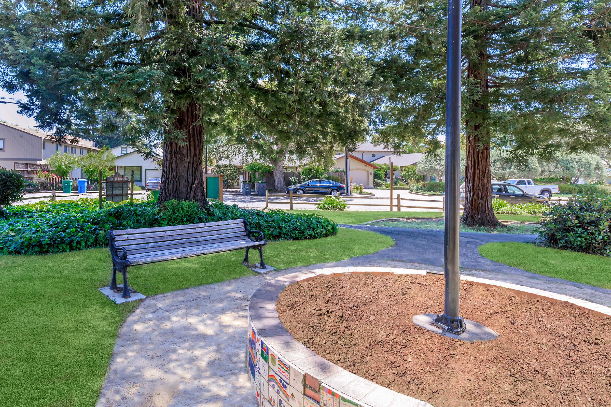 A peaceful park scene featuring a wooden bench surrounded by green grass and shrubs. A circular flower bed with a flagpole is in the foreground, while trees provide shade. In the background, residential houses and parked cars are visible, creating a serene community setting.