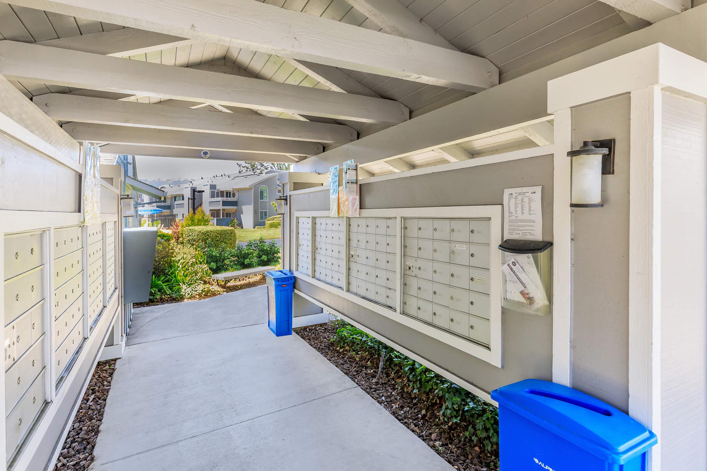 A covered area featuring multiple mailboxes against a gray wall, with two blue recycling bins on either side. Surrounding greenery and a walkway lead to a residential area in the background, partially visible through the open space. The setting is well-lit and neatly maintained.