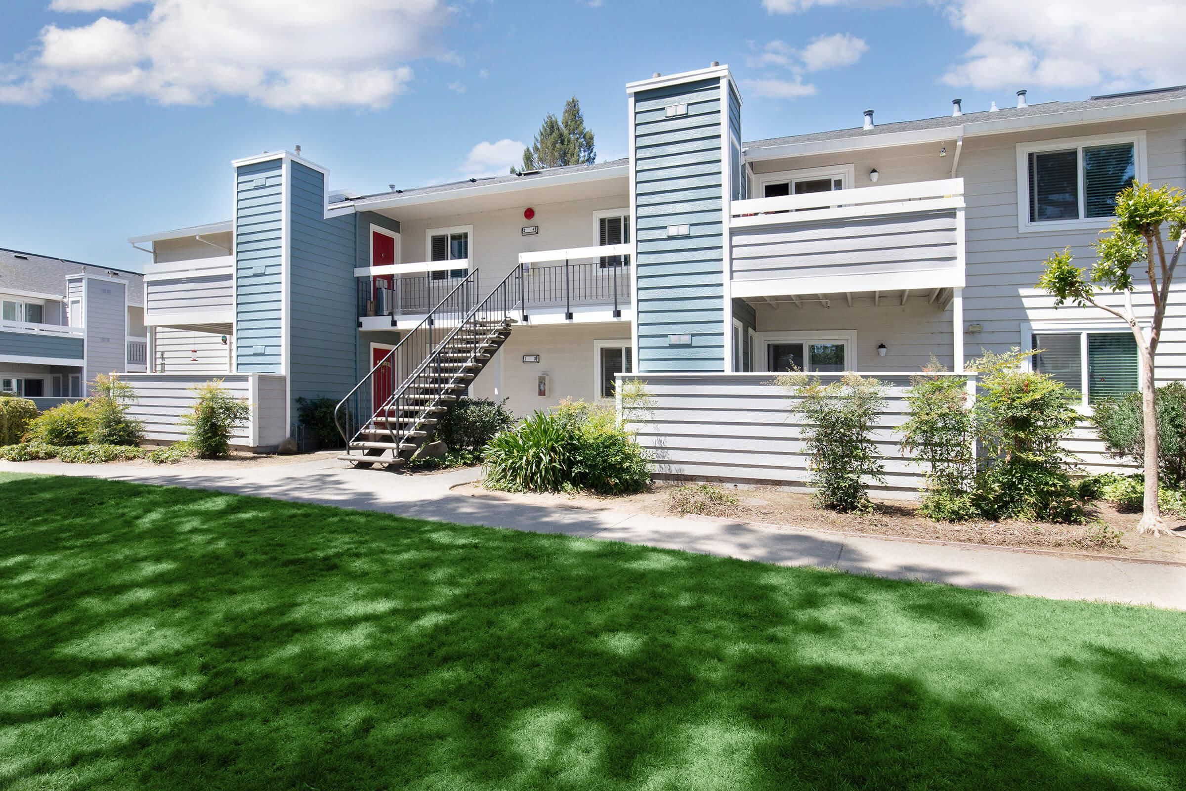 A modern, two-story apartment building with light blue and white siding. The building features a red door, multiple balconies, and stairs leading to the upper level. The surrounding area includes well-maintained green grass and bushes, under a partially cloudy sky.