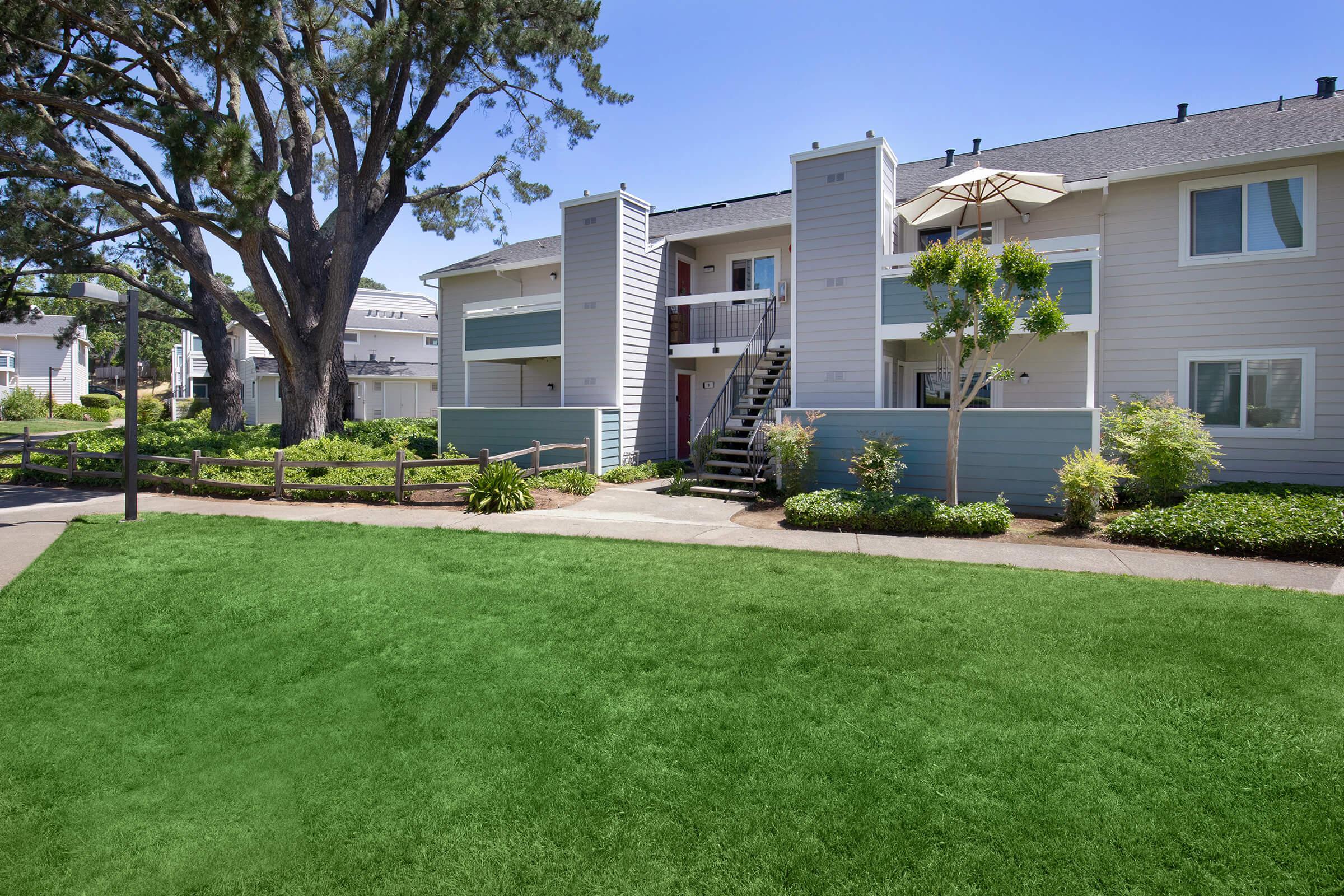 A well-maintained apartment complex featuring a two-story building with a red door, surrounded by lush greenery and a manicured lawn. A large tree provides shade, and there's an umbrella on the balcony, suggesting a pleasant outdoor space. The pathway leads to a scenic entryway.