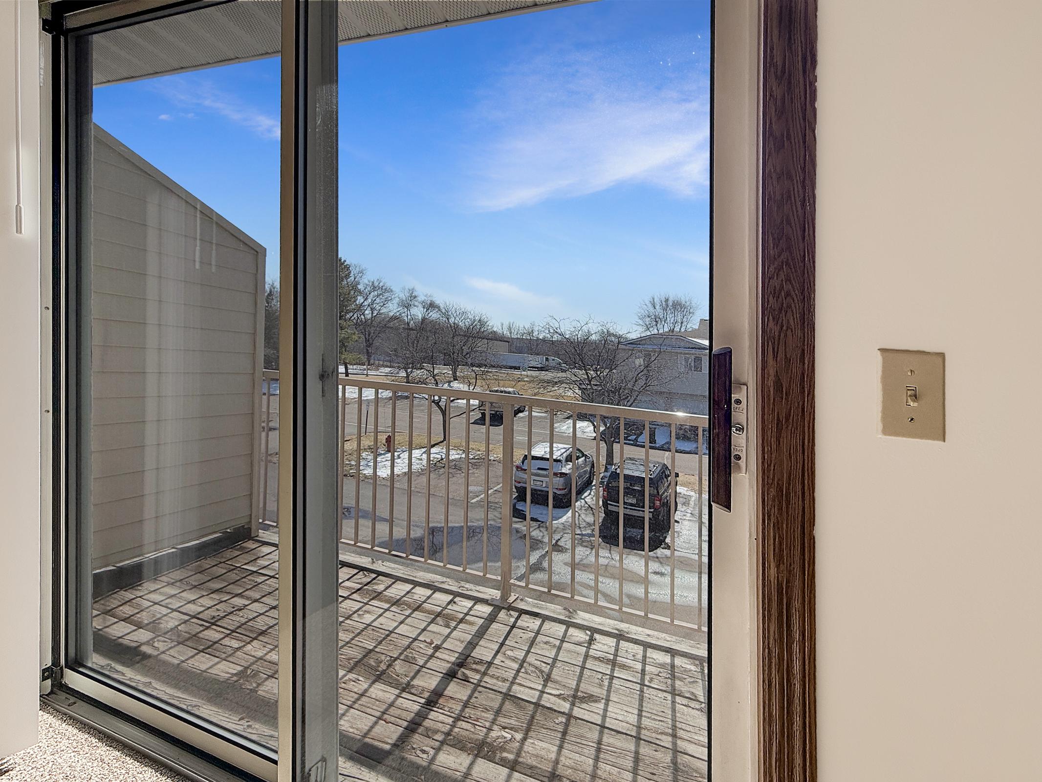 View from an interior doorway leading to a balcony, featuring sliding glass doors. The scene outside shows a clear blue sky with a few clouds, and a parking area with cars visible below. The balcony has a simple railing and appears to be part of a multi-unit residential building.