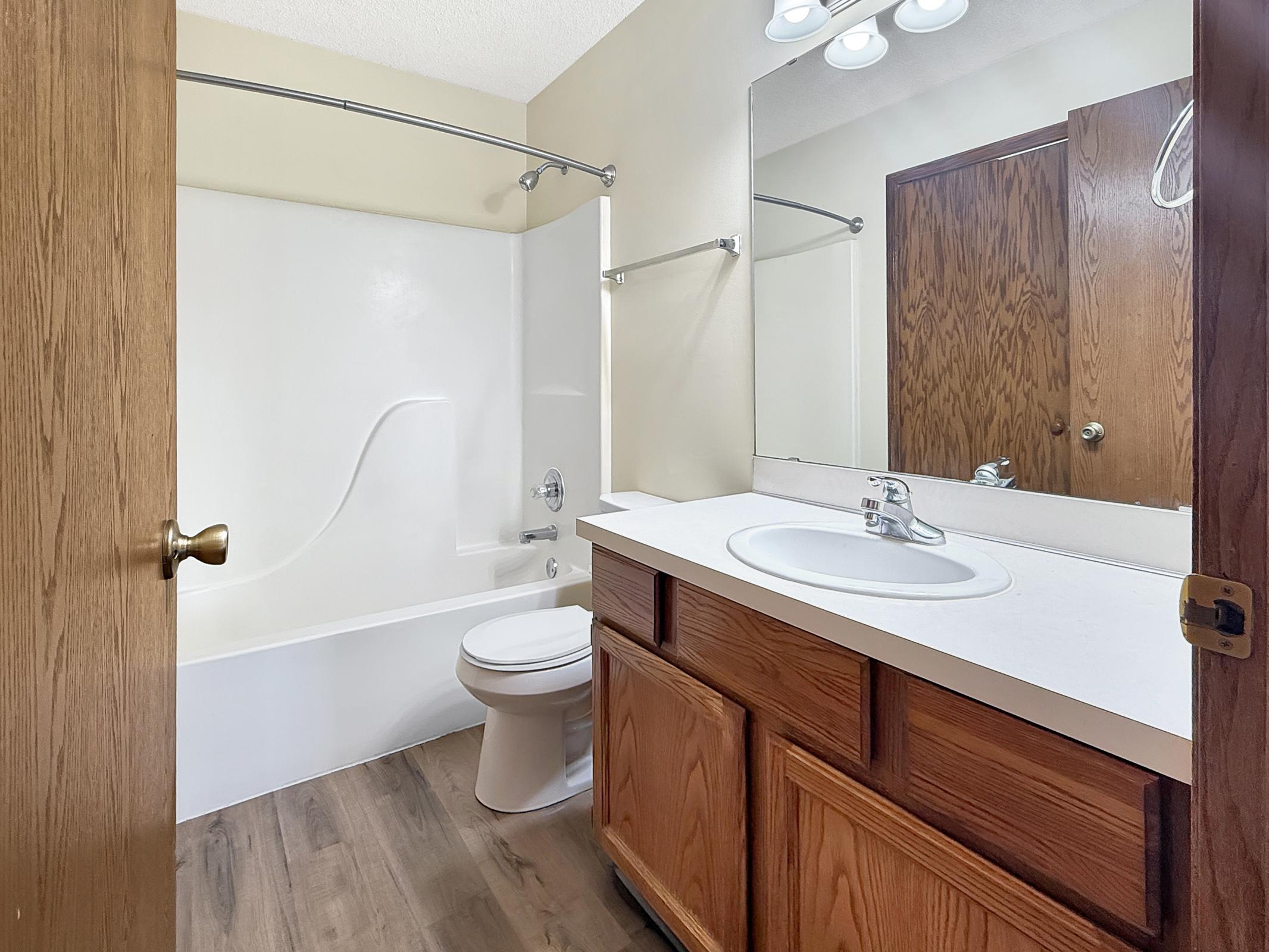A view of a small bathroom featuring a white bathtub, toilet, and a vanity with a sink. The walls are painted light beige, and there is a large mirror above the sink. The bathroom has wooden cabinets and a simple, clean design. Natural light is present, enhancing the spacious feel.