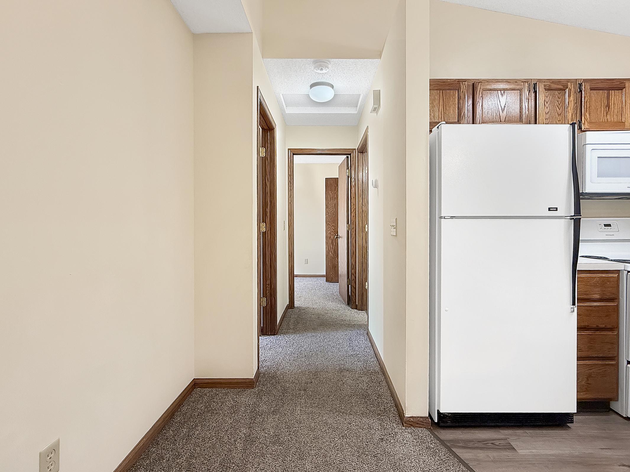 A hallway view featuring beige walls and carpet, leading to doors on either side. At the end of the hallway, there is a refrigerator and kitchen appliances visible. The lighting is bright, and the space appears clean and well-maintained.
