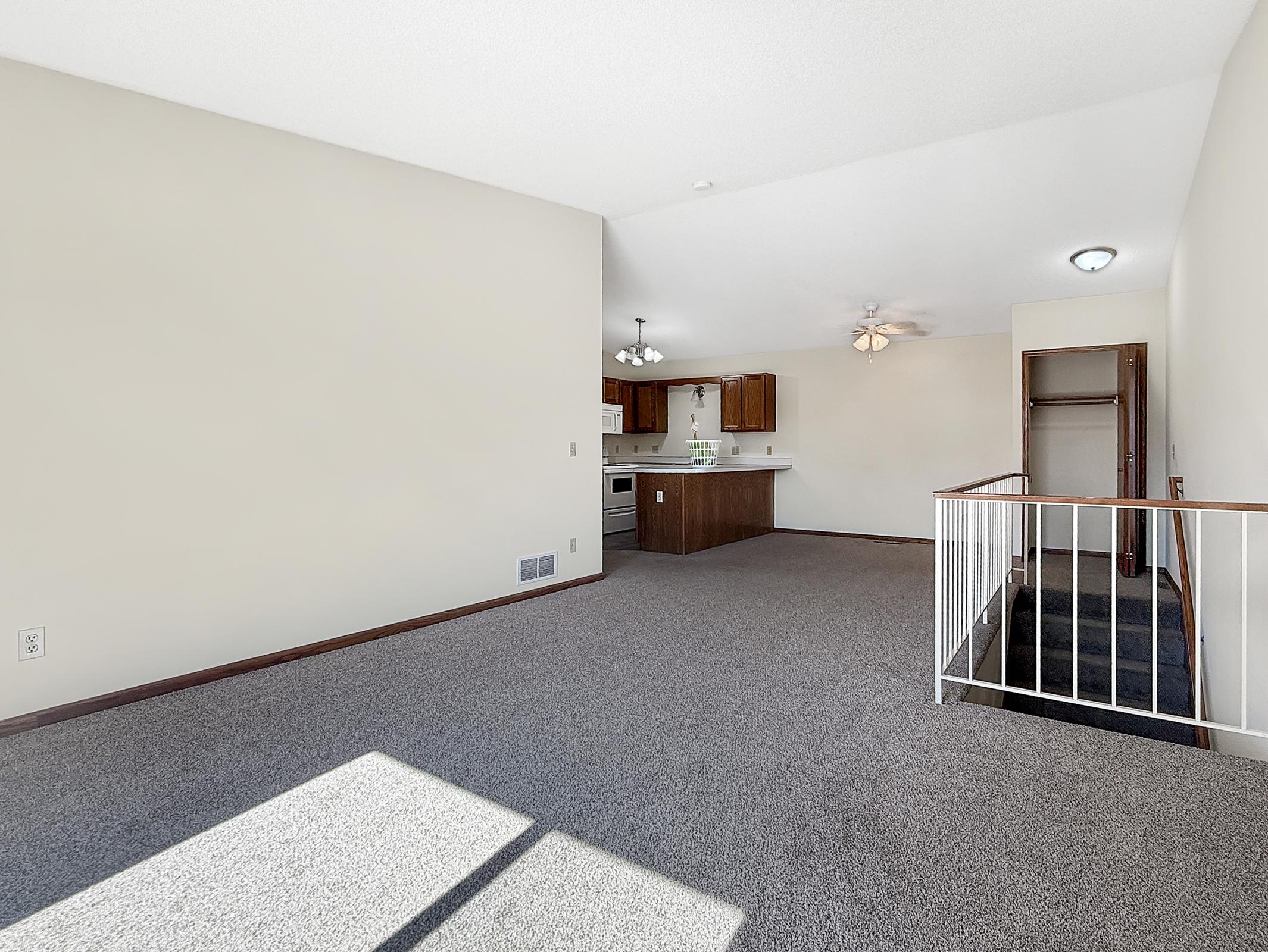 A spacious living area with gray carpet, featuring high ceilings and light-colored walls. The kitchen is visible in the background, equipped with wooden cabinets and appliances. A staircase leads to an upper level, and a small closet is on the right side. Natural light enters from a window, creating a welcoming atmosphere.