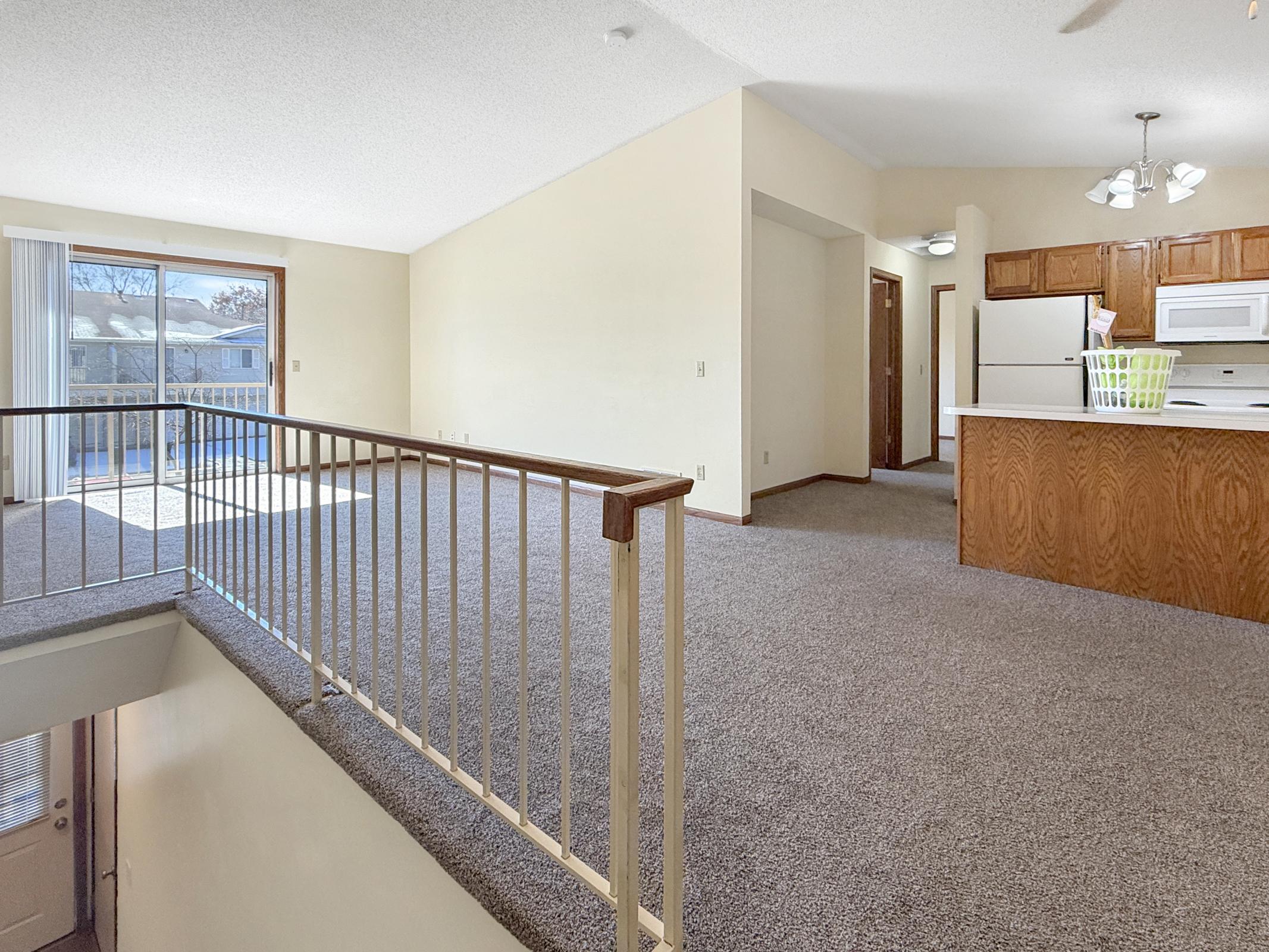 Spacious living area with carpet flooring, featuring a balcony, and an open kitchen view with wooden cabinets. The layout includes natural light from large windows and a ceiling fan, creating a bright and inviting atmosphere.