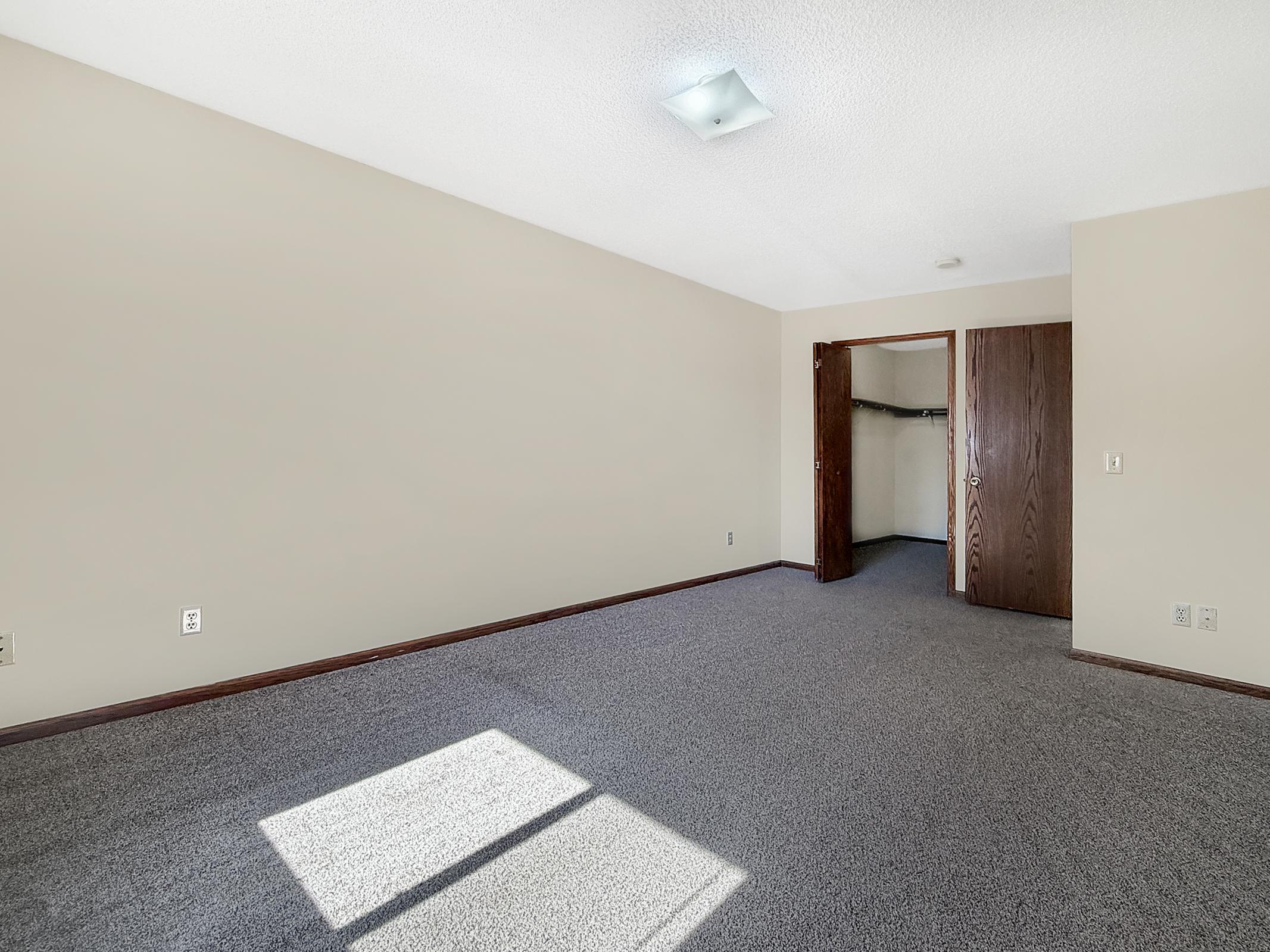 A spacious, empty room with light beige walls and gray carpet. The room features a ceiling light, a closet with an open door on the right, and a large window allowing natural light to enter, creating a bright area in the corner. The overall atmosphere is clean and inviting.