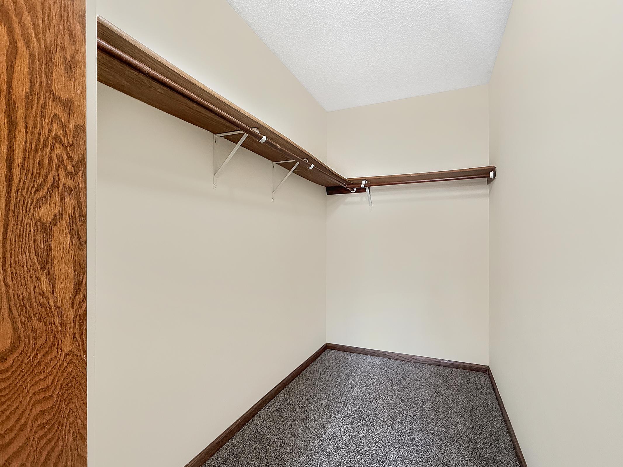 A spacious, empty closet with a high ceiling. It features two wooden shelves mounted on the walls and carpeted flooring. The walls are painted a light beige color, creating a bright and open appearance. The overall space is clean and uncluttered, ideal for storage or organization.