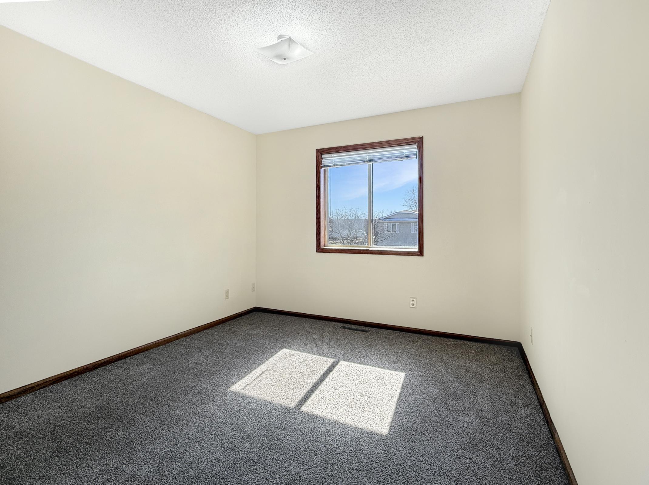 Empty room with light beige walls and a large window allowing sunlight to stream in. The floor is covered with a plush gray carpet, and there is a small ceiling fixture. The overall atmosphere is bright and inviting, suggesting a fresh, clean space ready for personalization.