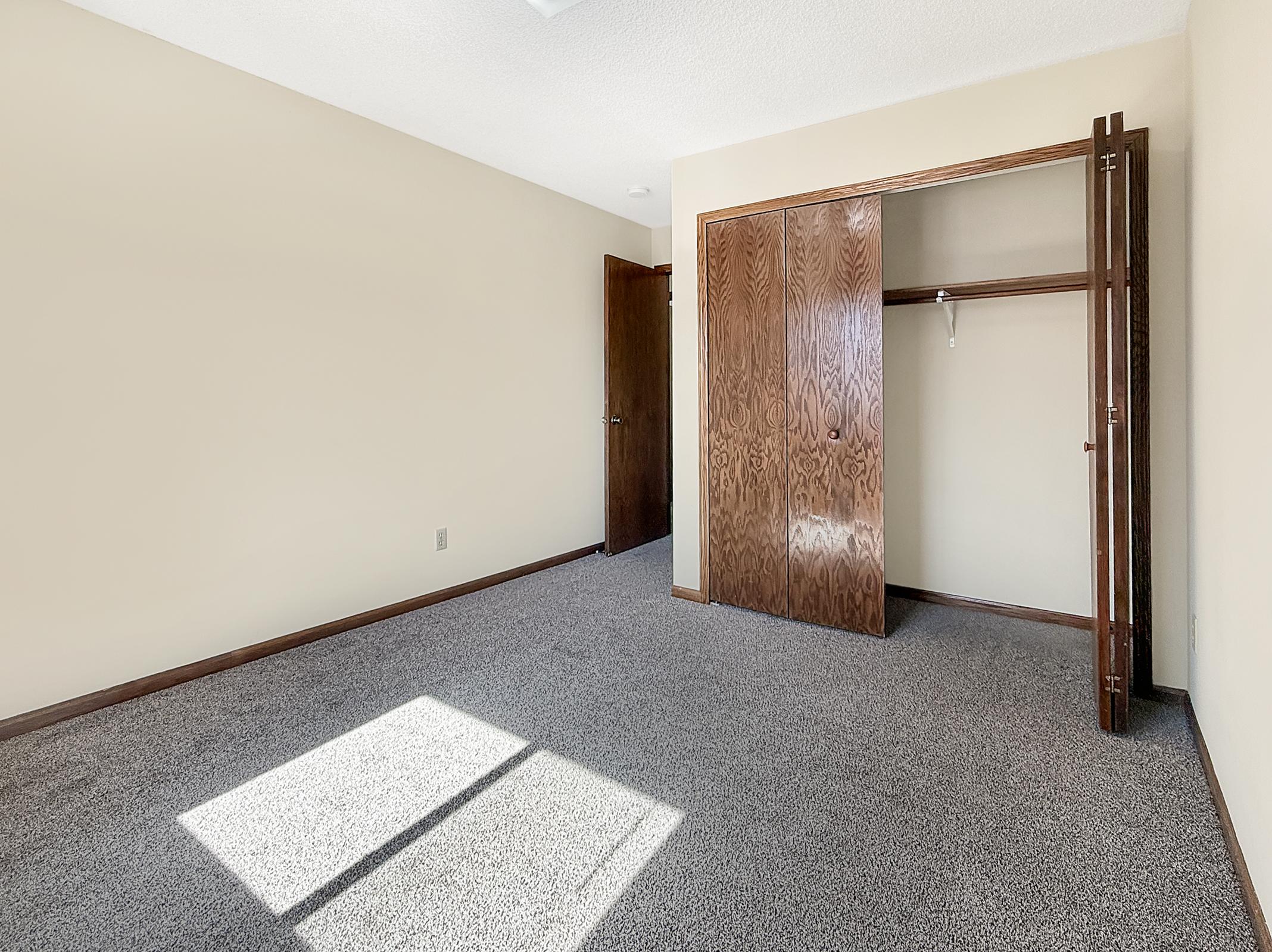 Empty room with beige walls and gray carpet. A large window allows natural light to flood in, creating a bright spot on the floor. There's a closed wooden closet with a door on the left and a dark wood door leading to another area opposite the closet. The room appears spacious and uncluttered.